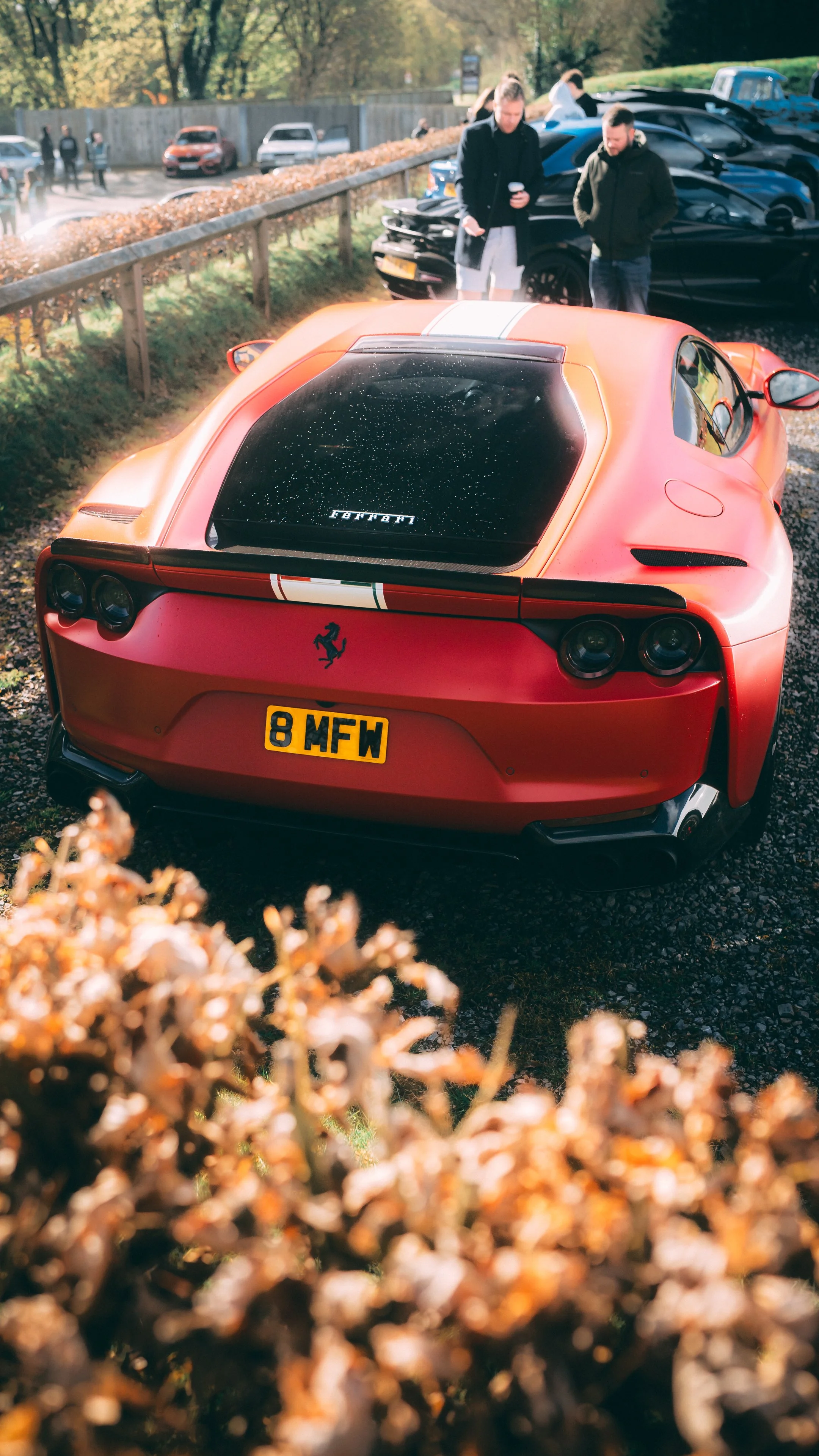 A red Ferrari sports car parked outdoors with a rear view, surrounded by people and other cars in the background, during daytime with autumn leaves.