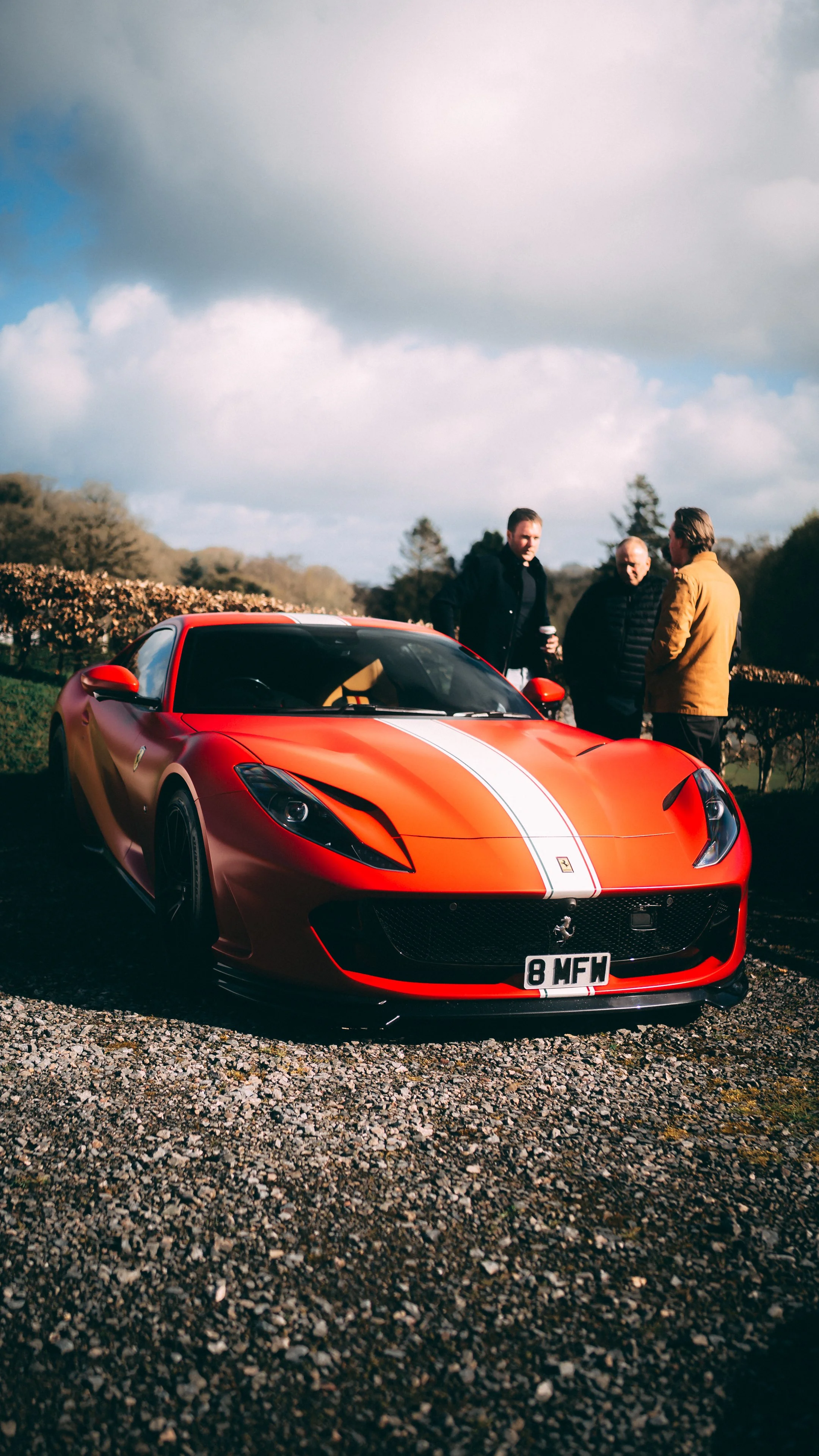 A red Ferrari sports car with white racing stripes parked on a gravel surface, with three men standing nearby engaged in conversation outdoors under a partly cloudy sky.