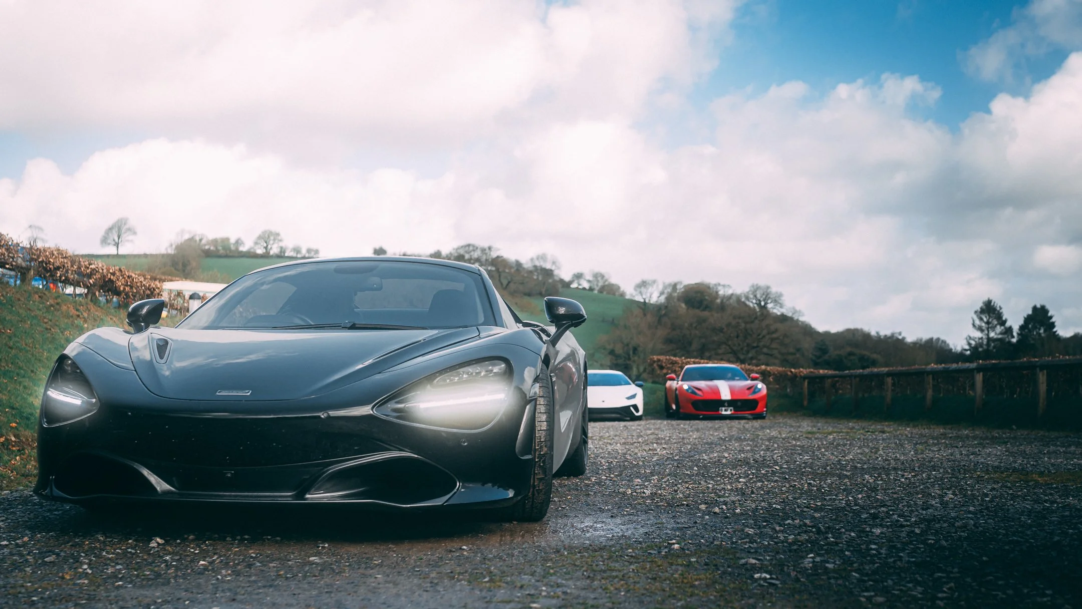A lineup of sports cars parked on a gravel surface outdoors with a cloudy sky and green hills in the background.