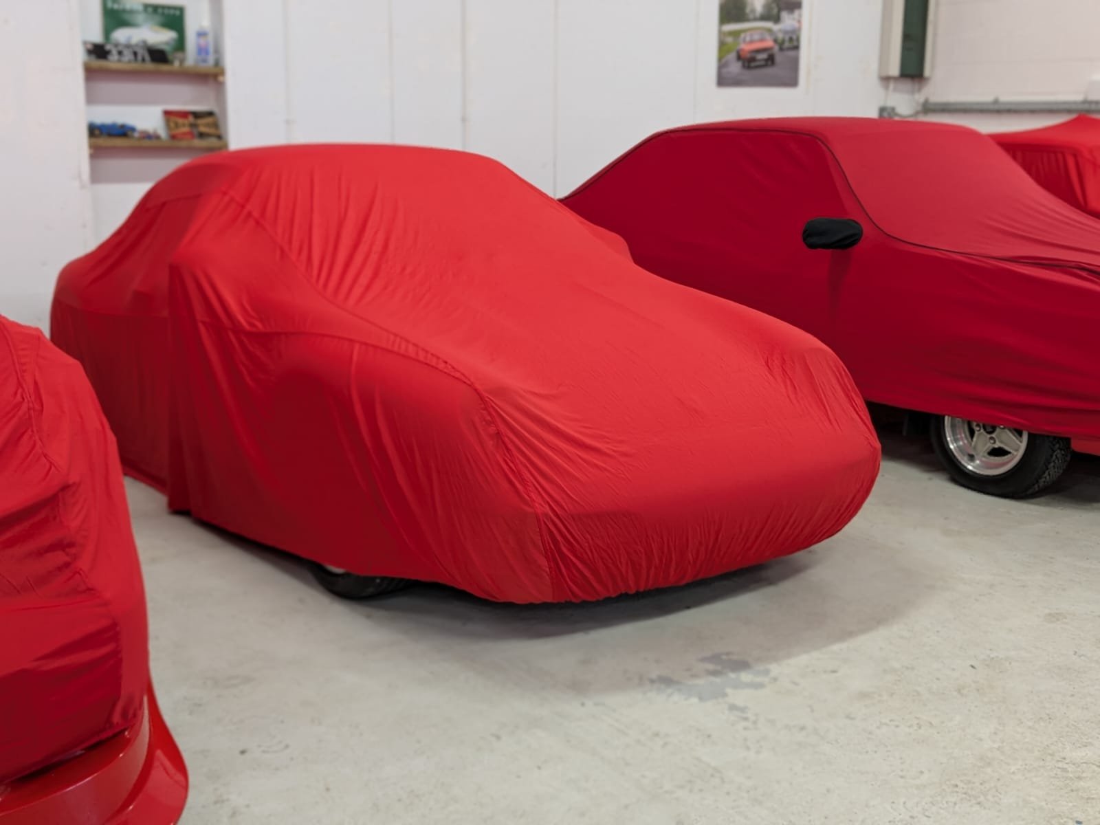 Three cars covered with red protective covers in a garage. The covers are snug, showing the shape of the vehicles underneath.