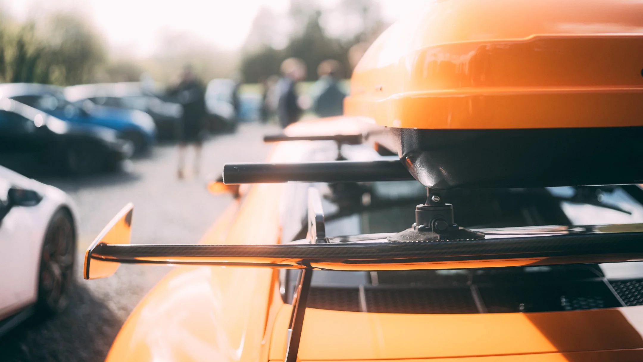 Close-up of a bright orange race car with a spoiler, with blurred cars and people in the background on a sunny day.
