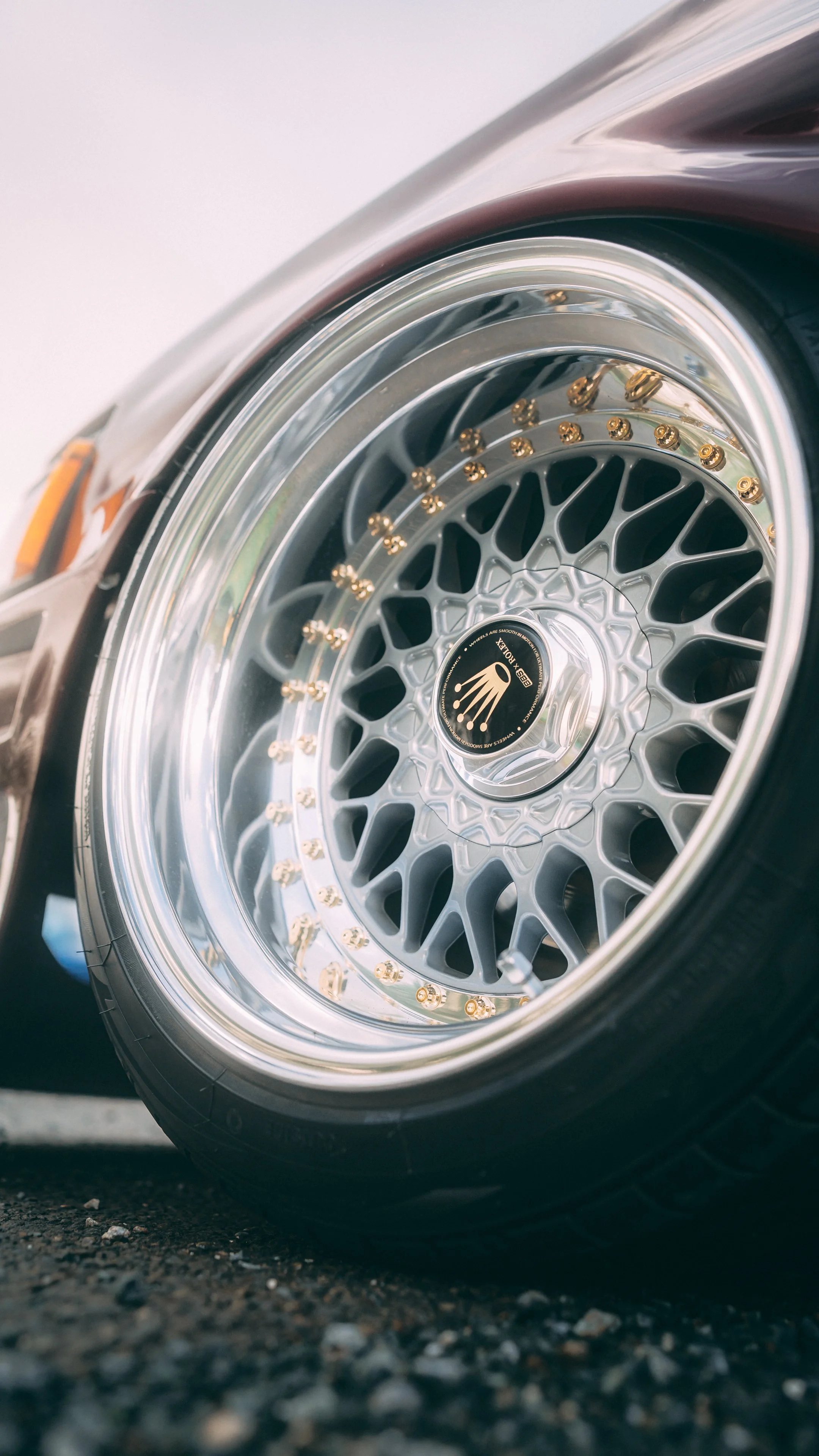 Close-up of a car's alloy wheel with gold-colored lug nuts and a center cap showing the Rolex logo, set on a gravel ground.