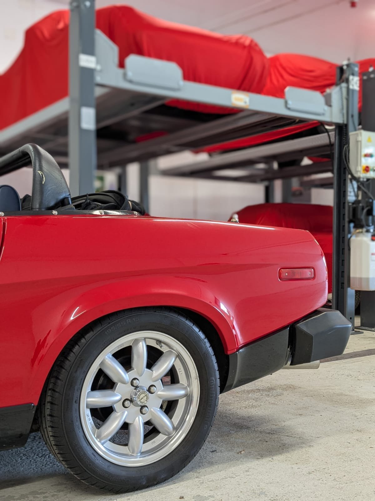Close-up of a red vintage sports car with a visible wheel, parked in an indoor garage with a metal bunk bed above it.