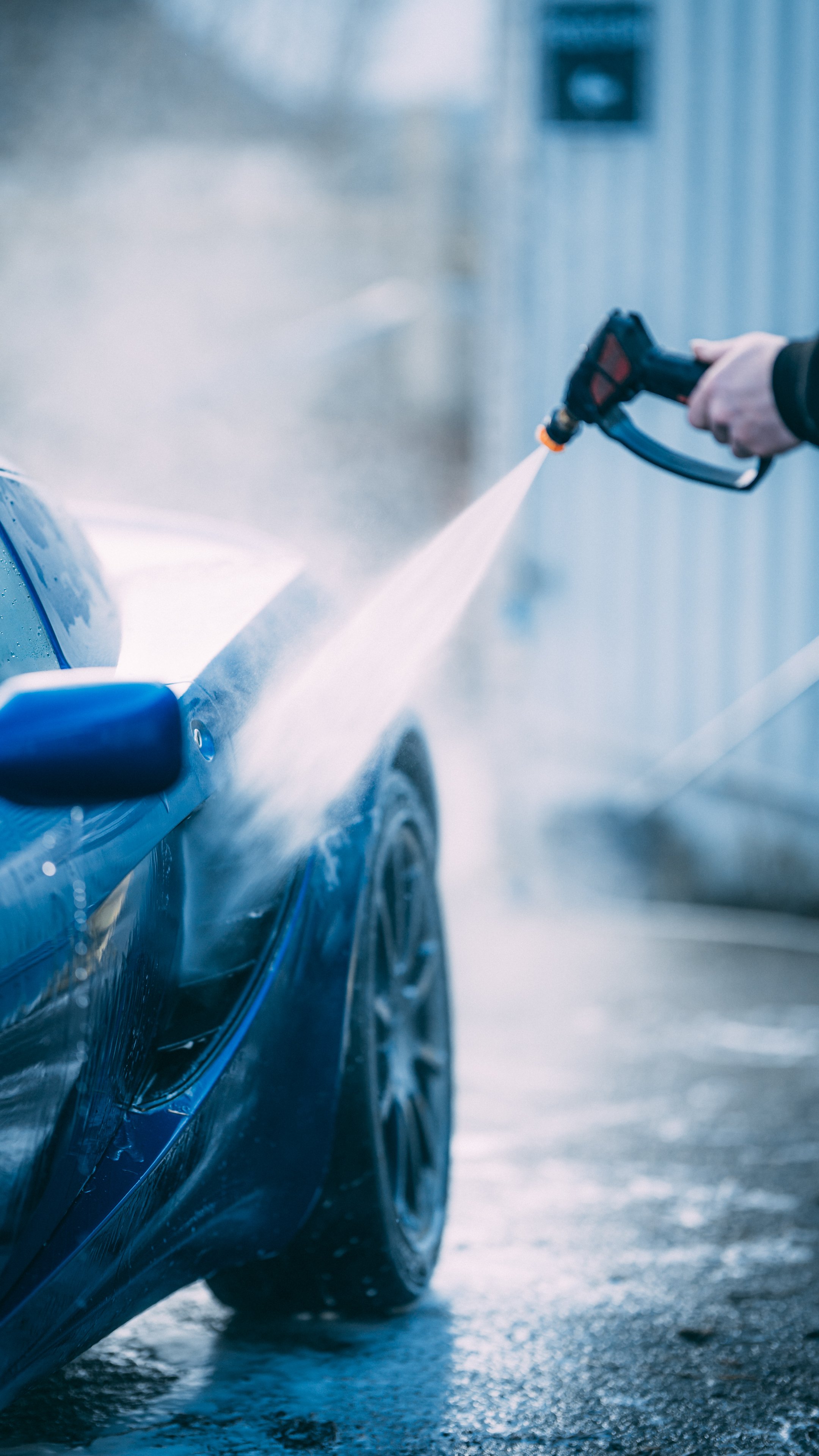 Person using a pressure washer to clean a blue sports car.