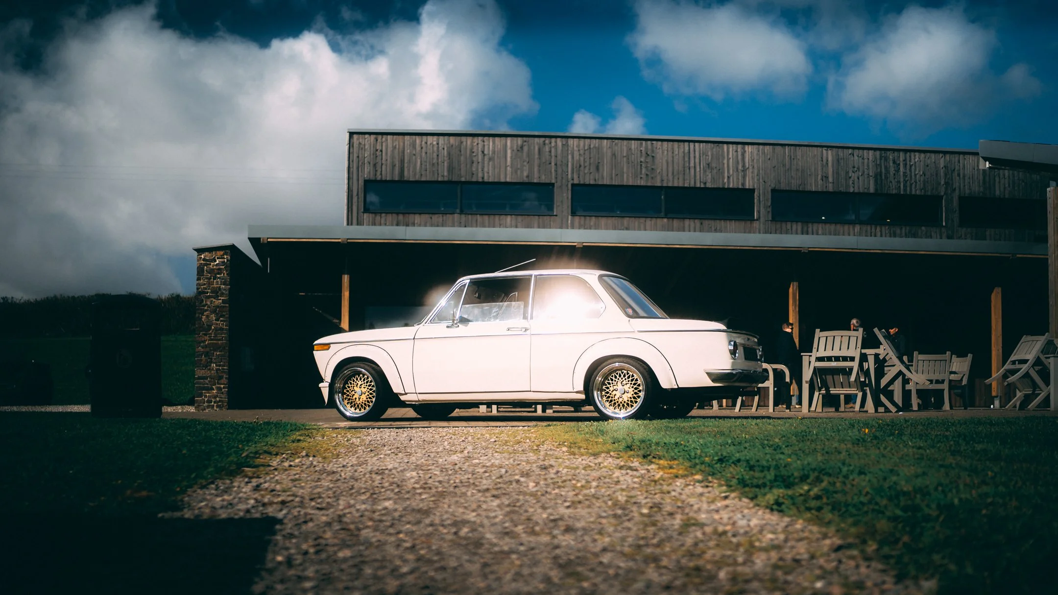 A white vintage car with gold wheels parked outside a modern house with large windows and wooden exterior, under a partly cloudy sky.