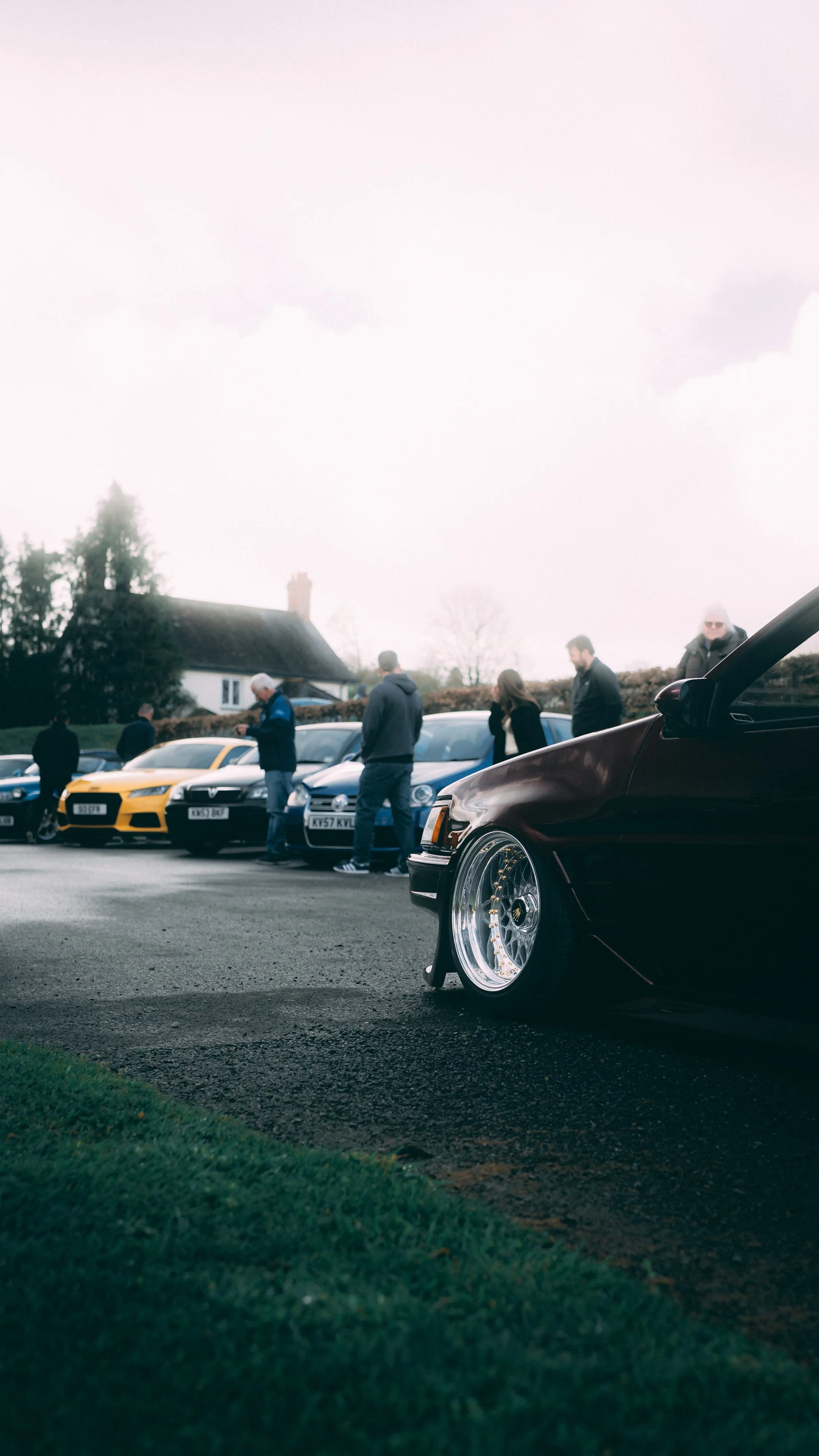 Line of luxury cars parked on a street with people looking at them, including a black car in the foreground with custom wheels, in a suburban neighborhood with houses and trees.