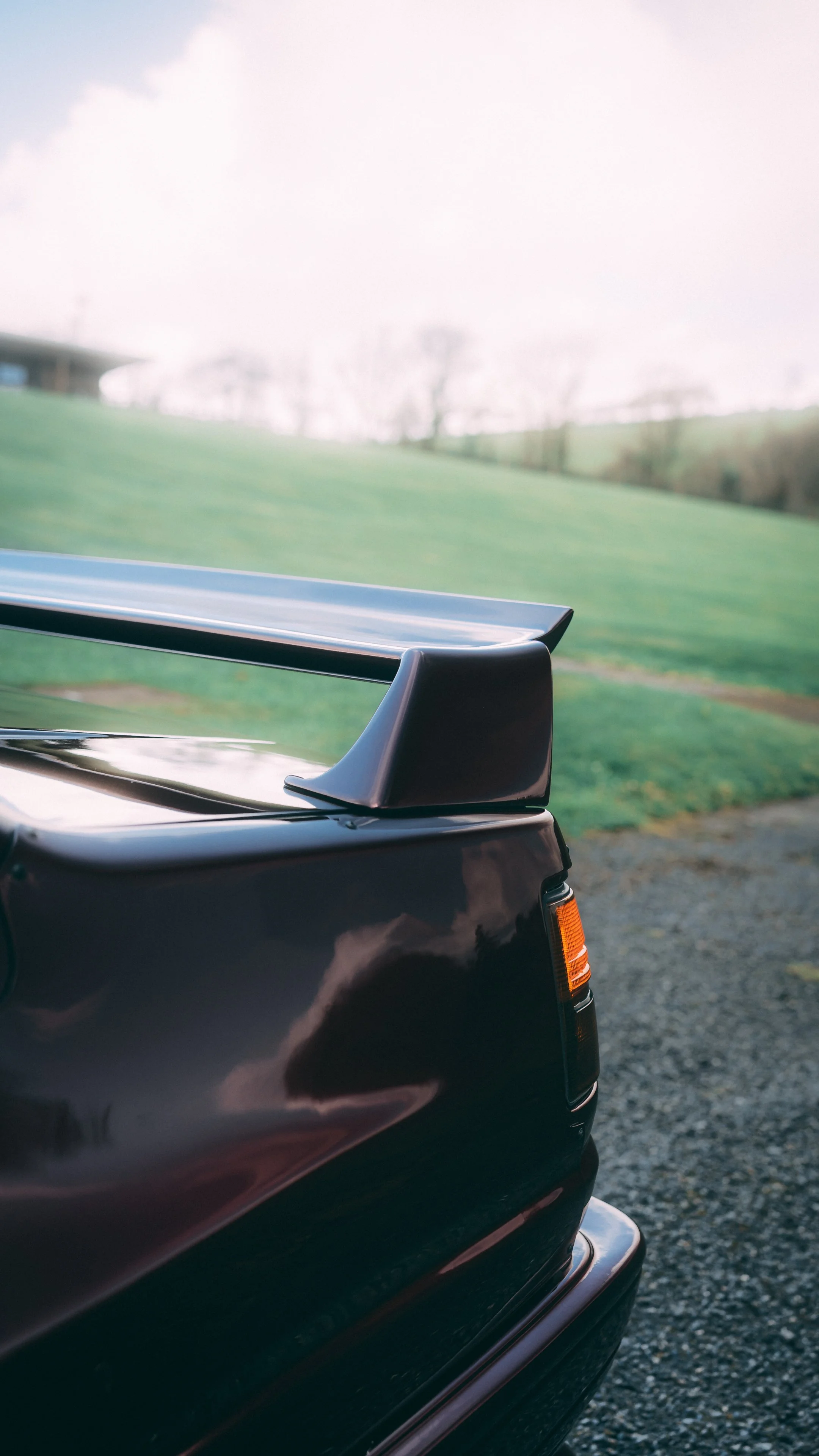Close-up of the rear part of a black vintage car with a black spoiler, parked on a gravel surface with green hills and leafless trees in the background.