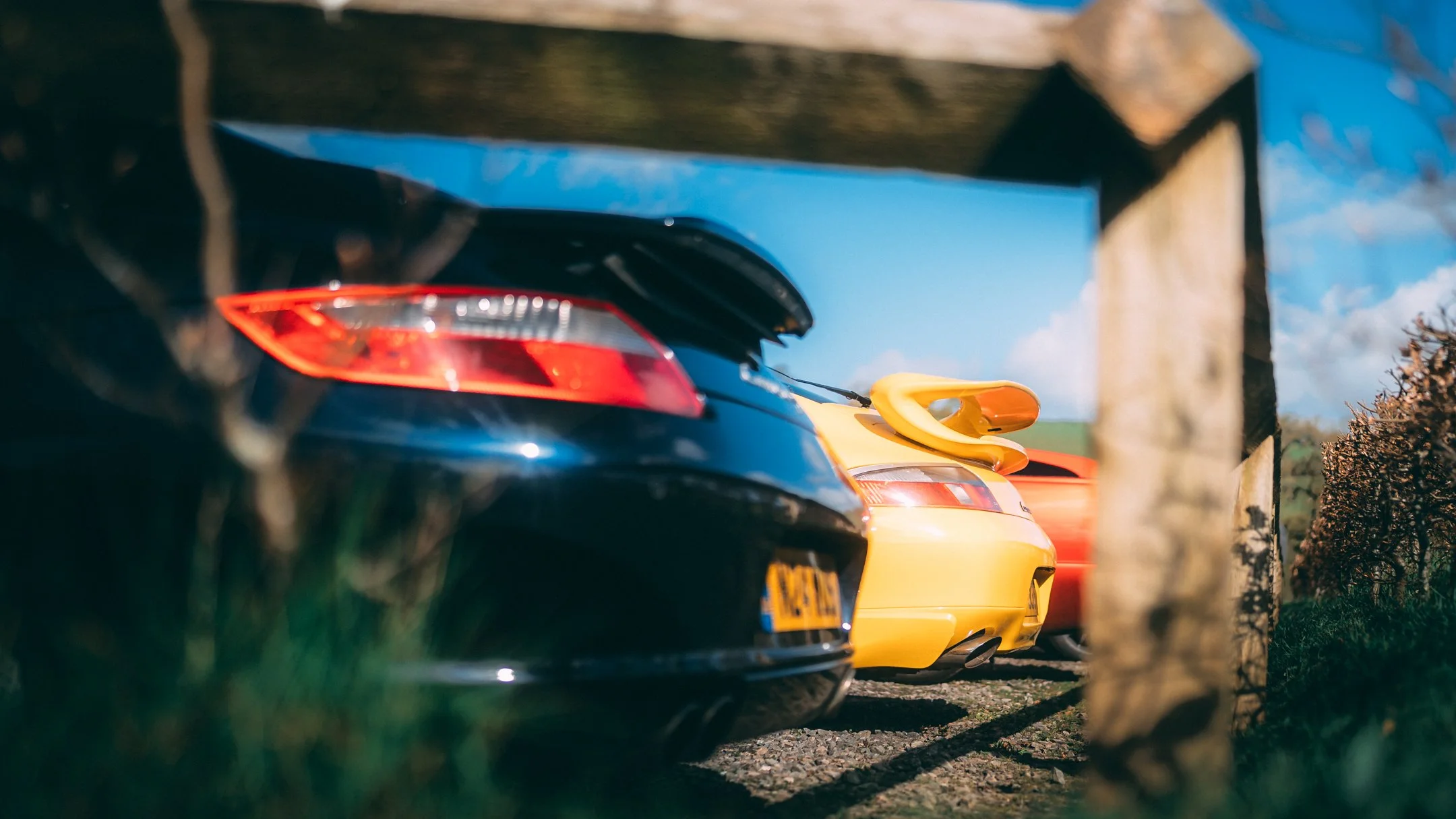 Multiple cars parked under a bridge or tunnel, including a black sports car, a yellow car, and a red car, with a rural landscape and a blue sky in the background.