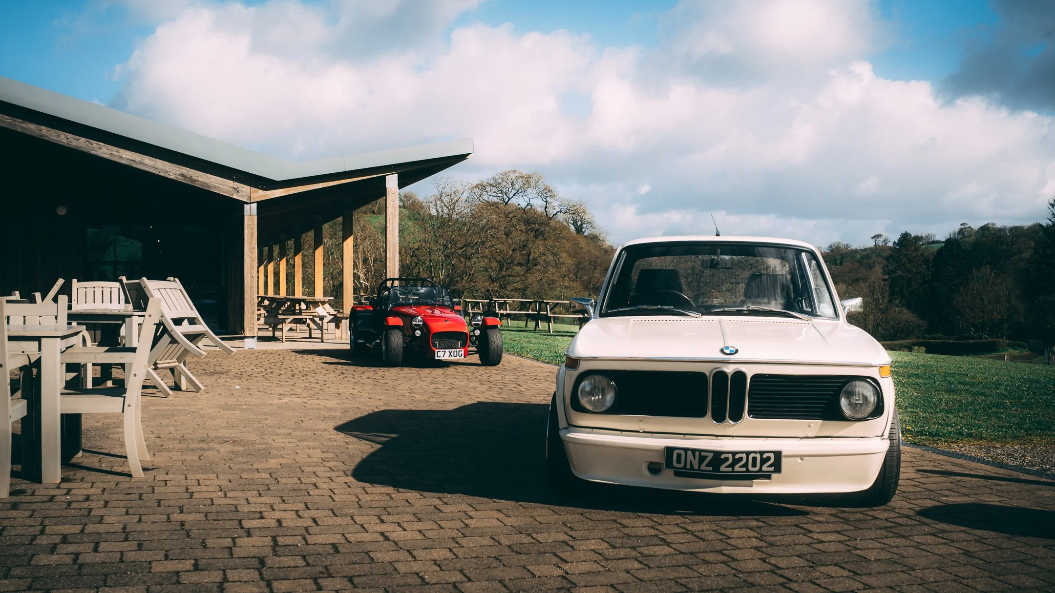A white vintage BMW car parked on a brick driveway, a red toy car and a black ride-on toy are nearby, with outdoor seating and trees in the background under a partly cloudy sky.