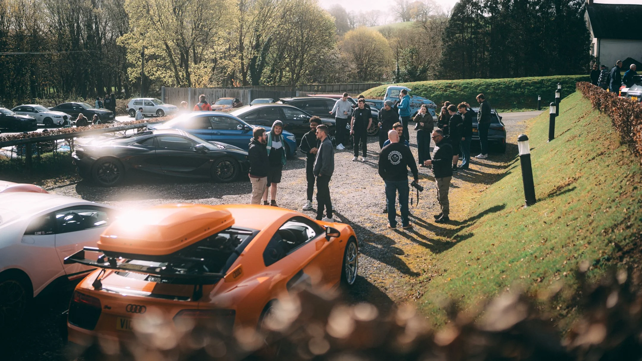 A group of people gathered outdoors near a parking lot with luxury cars, including an orange sports car in the foreground, during daytime with clear weather and trees in the background.