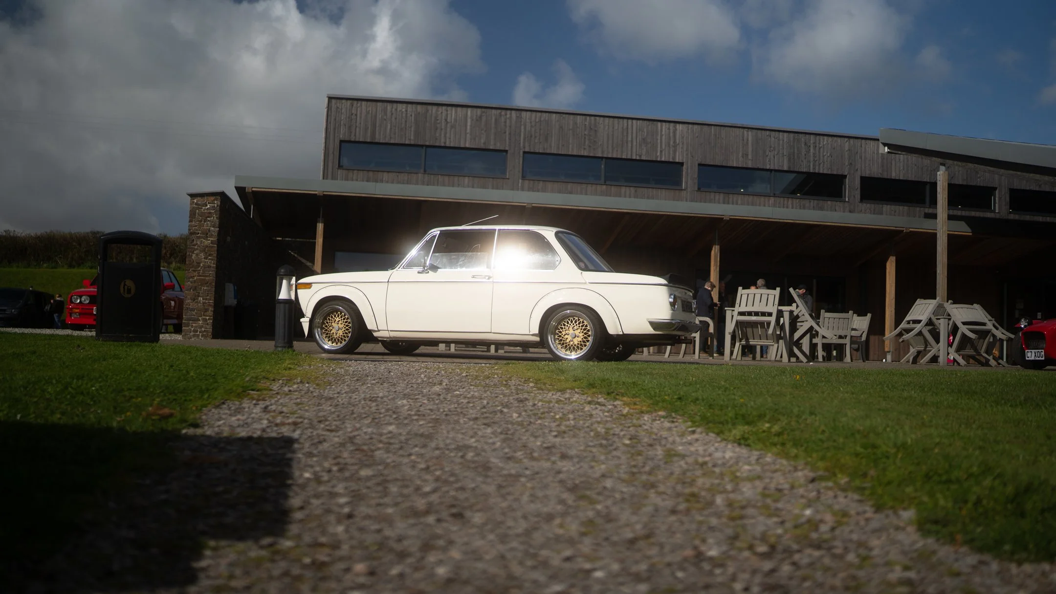 A white vintage car parked in front of a modern building with outdoor seating and a few people nearby on a partly cloudy day.