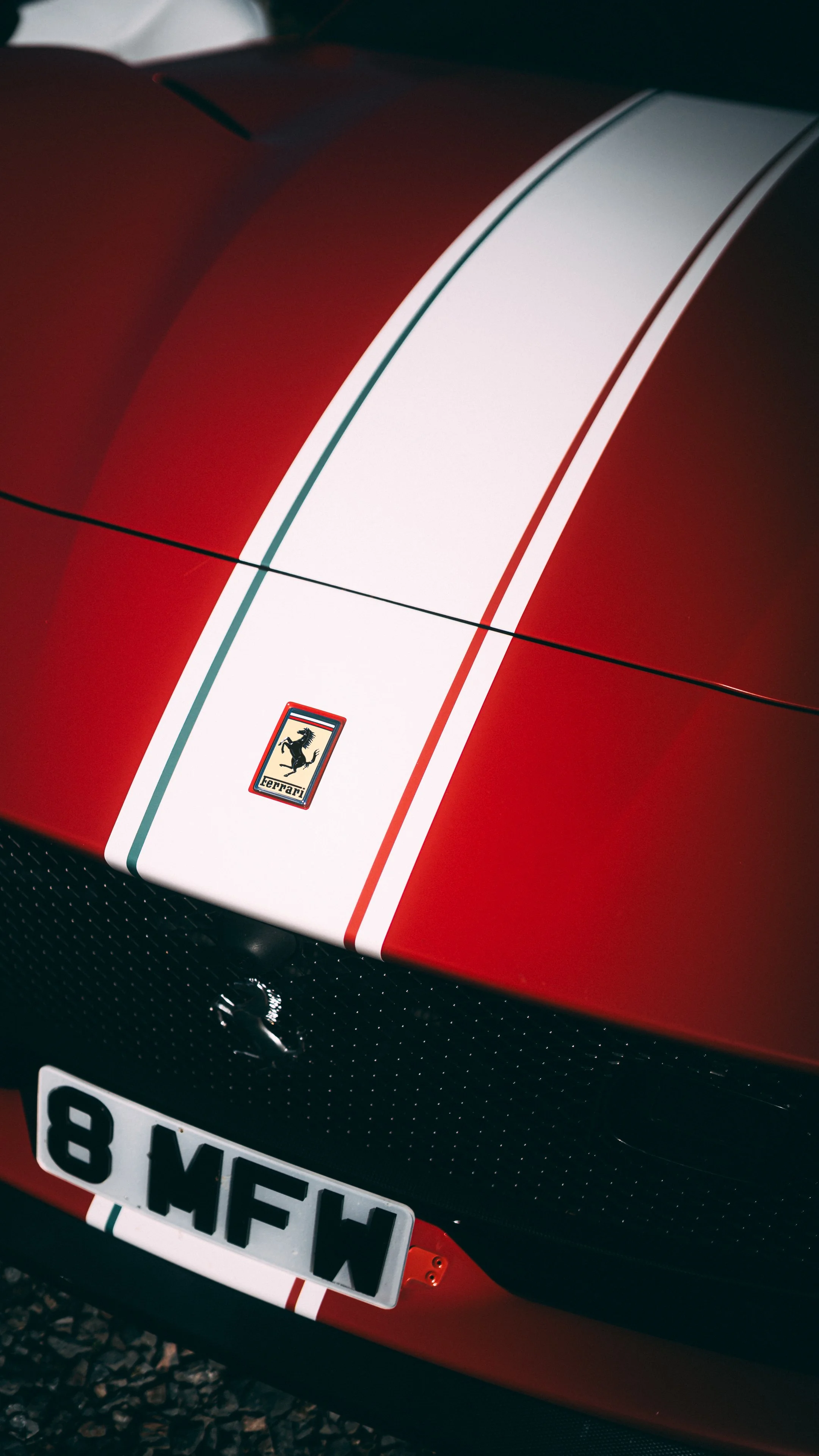 Close-up of a red sports car with a white racing stripe, Ferrari emblem, and partial European license plate.