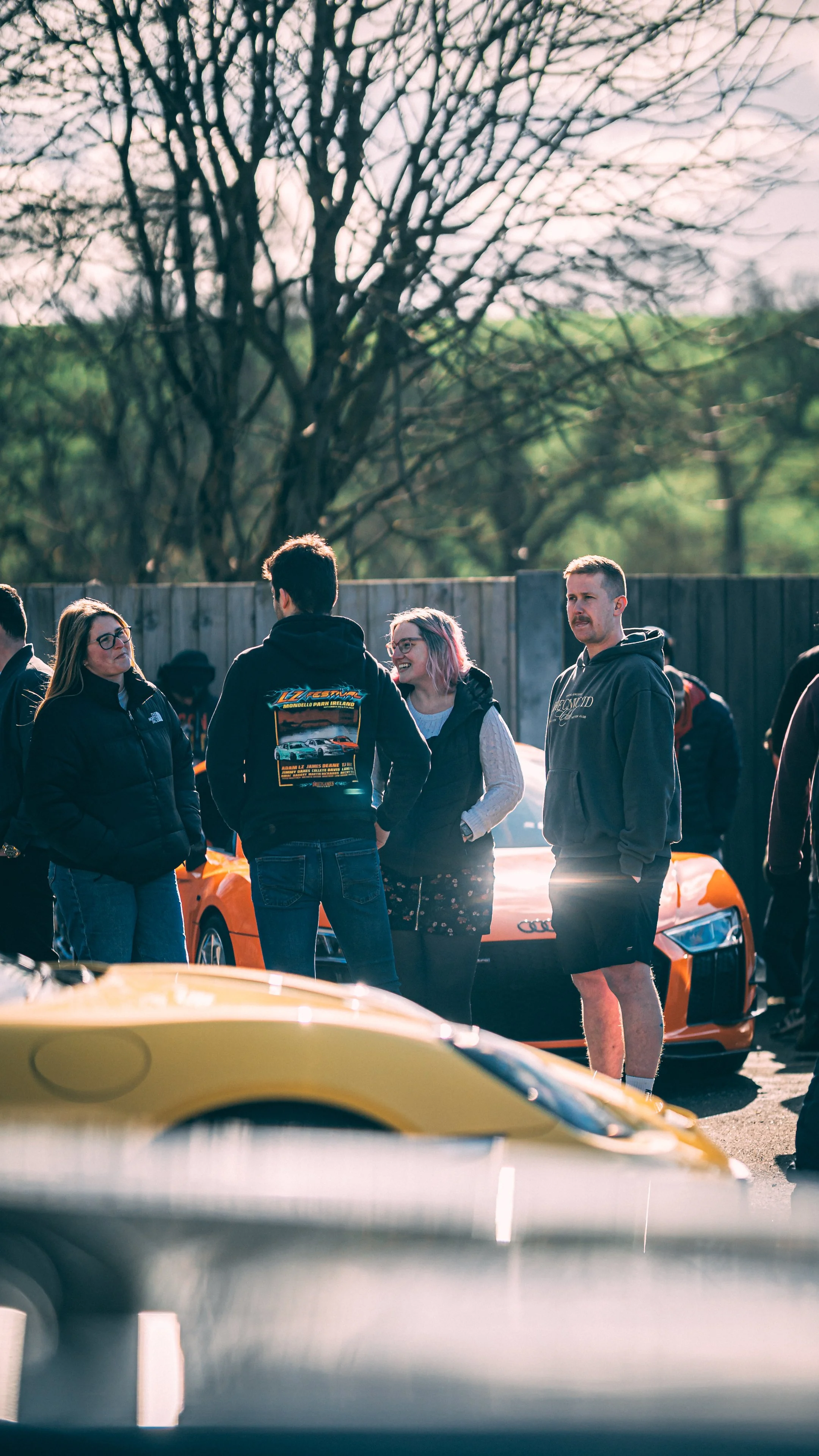 Group of people talking outdoors near a bright orange sports car, with trees and a wooden fence in the background, possibly at a car meet or show.