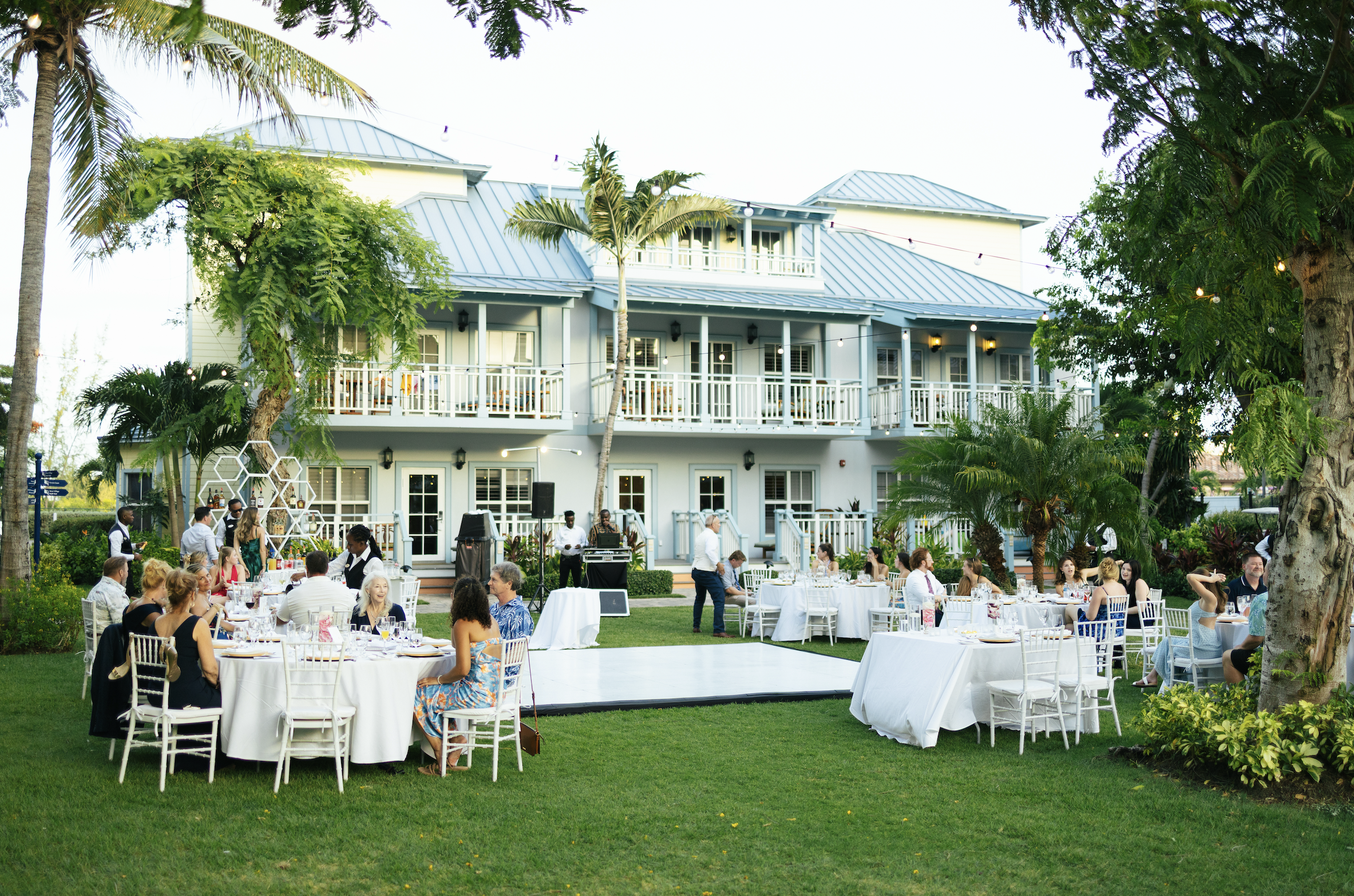 Outdoor wedding reception with guests seated at round tables on a lawn, a DJ booth, and a large white house with balconies and lush greenery in the background.