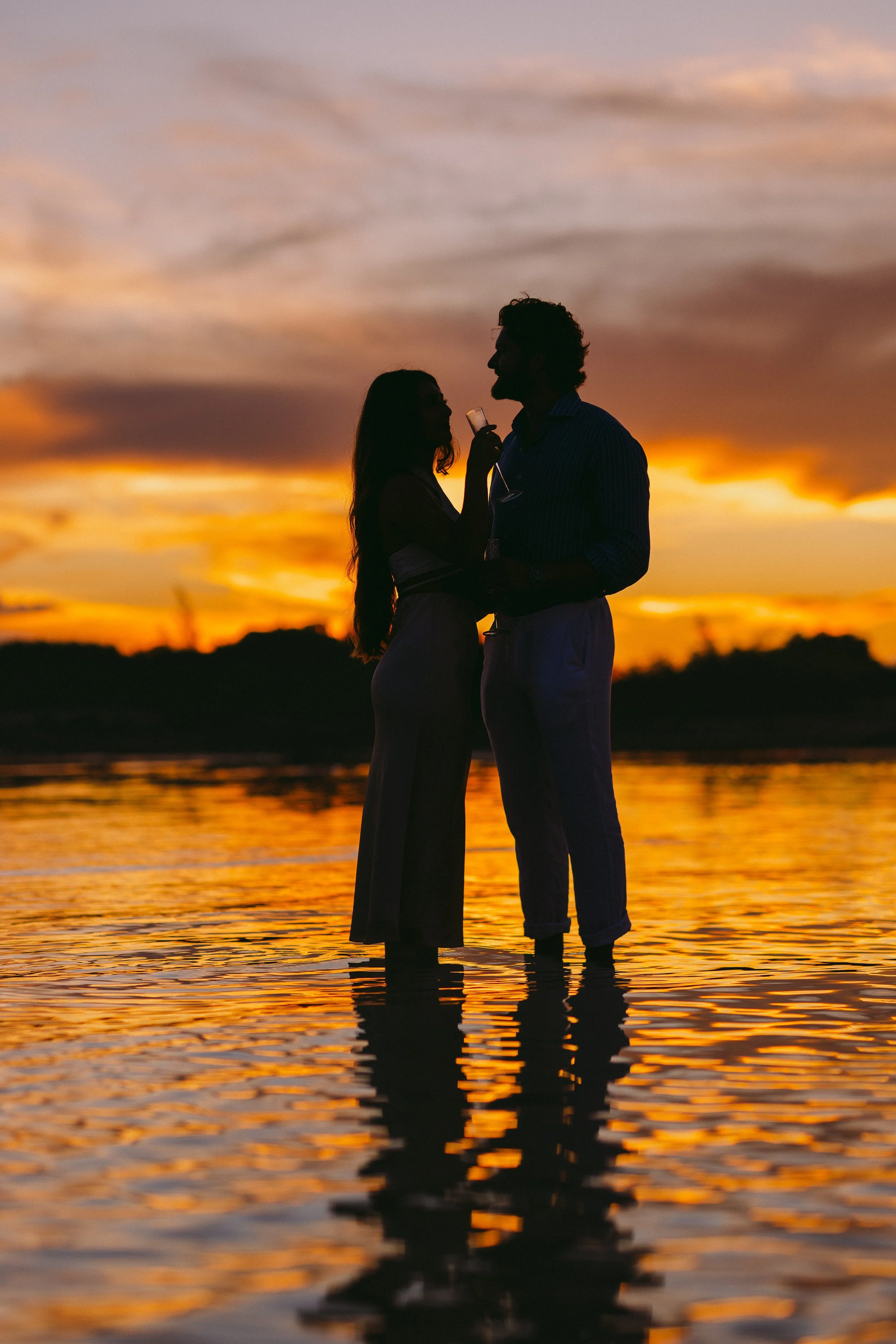 Silhouettes of a man and woman standing in water at sunset with a colorful sky reflected on the surface.