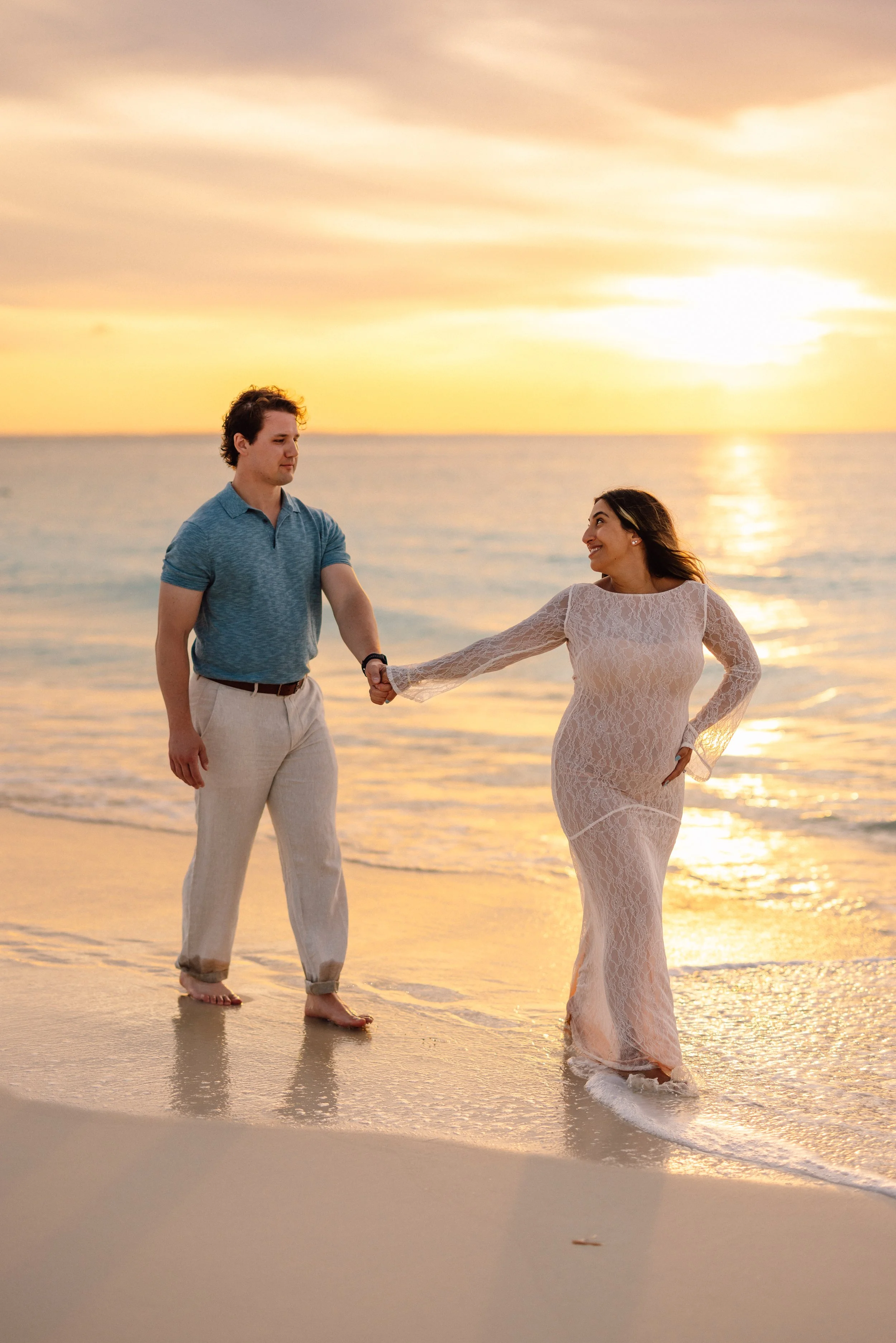 A man and woman hold hands on the beach during sunset, with the woman smiling and wearing a lace dress, and the man in casual attire, both barefoot in the shallow water.