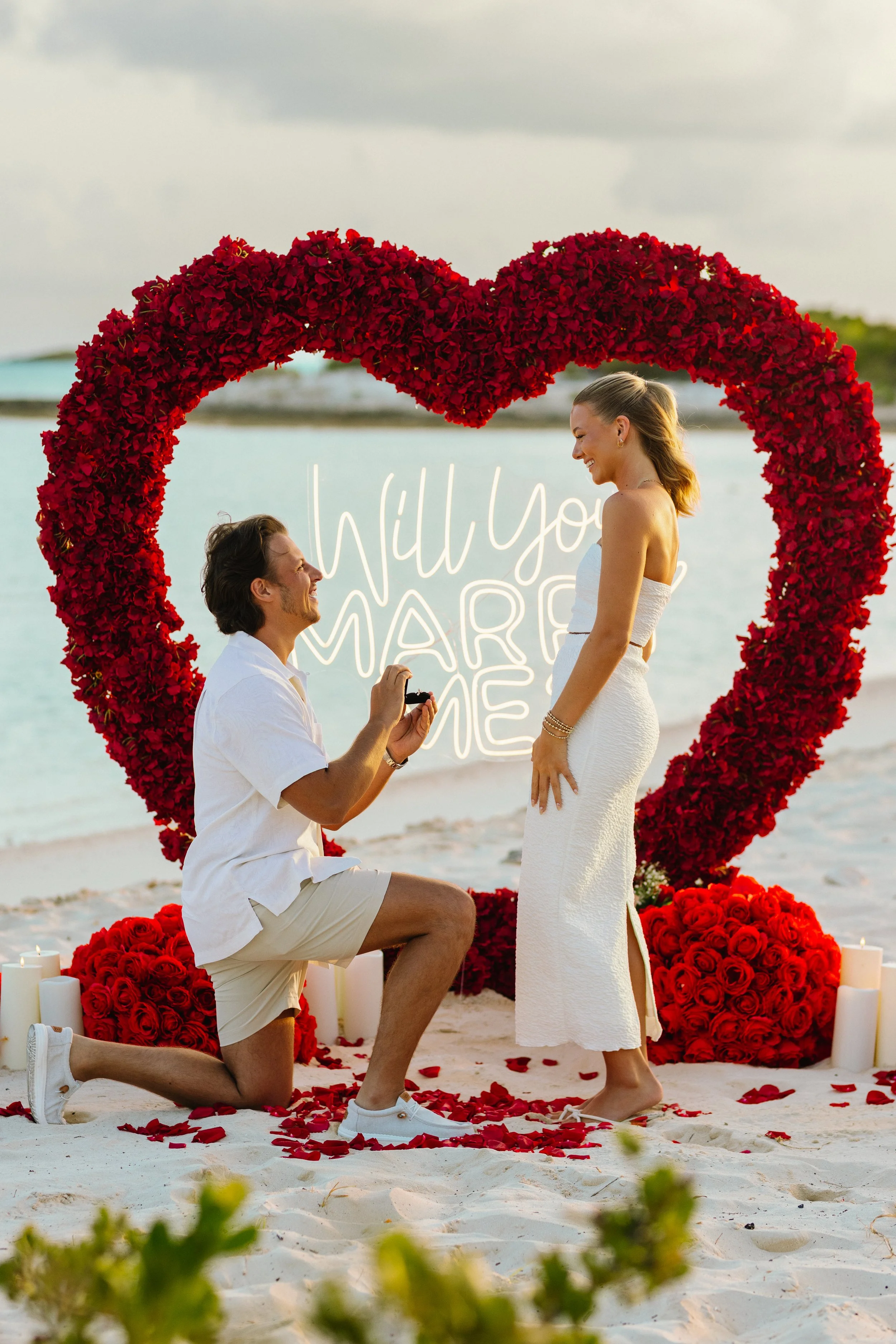 A man proposing marriage to a woman on the beach, surrounded by heart-shaped floral arrangement and candles, with water and sky in the background.