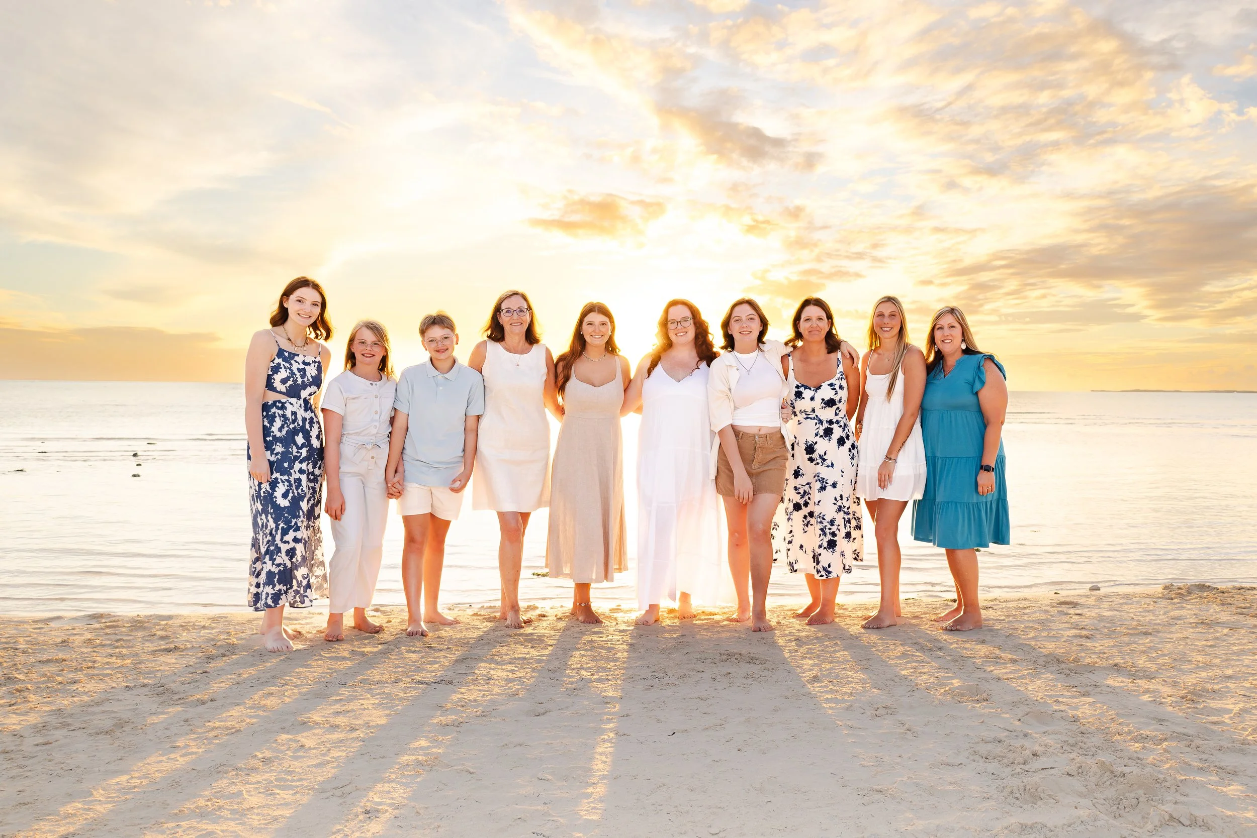 A group of ten women and children standing arm in arm on a sandy beach during sunset with a cloudy sky in the background, dressed in casual summer clothing.