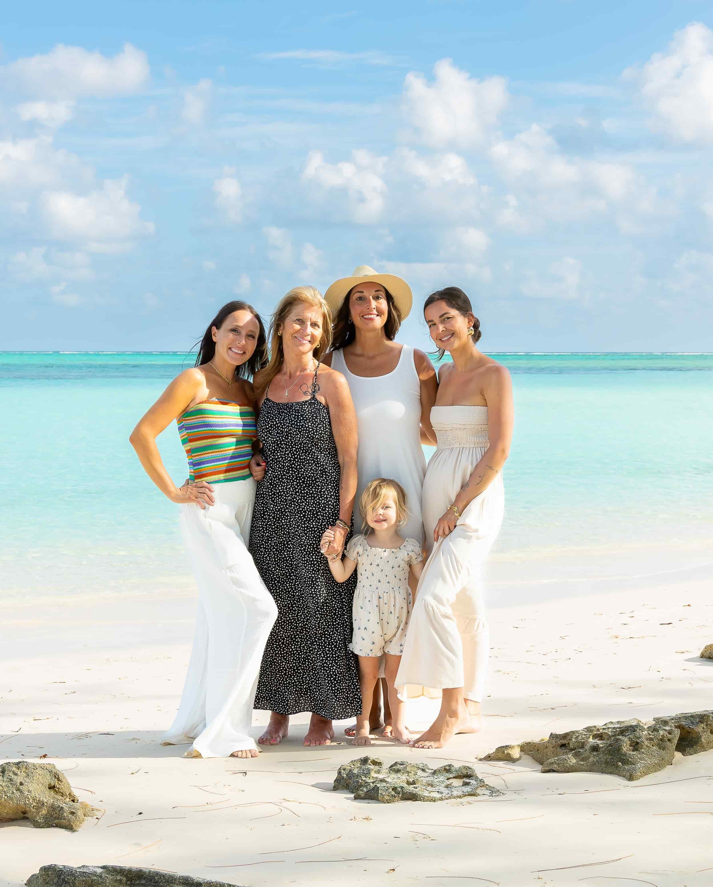 Group of five women and a young girl standing on a sandy beach with rocks, turquoise ocean, and partly cloudy sky in the background.