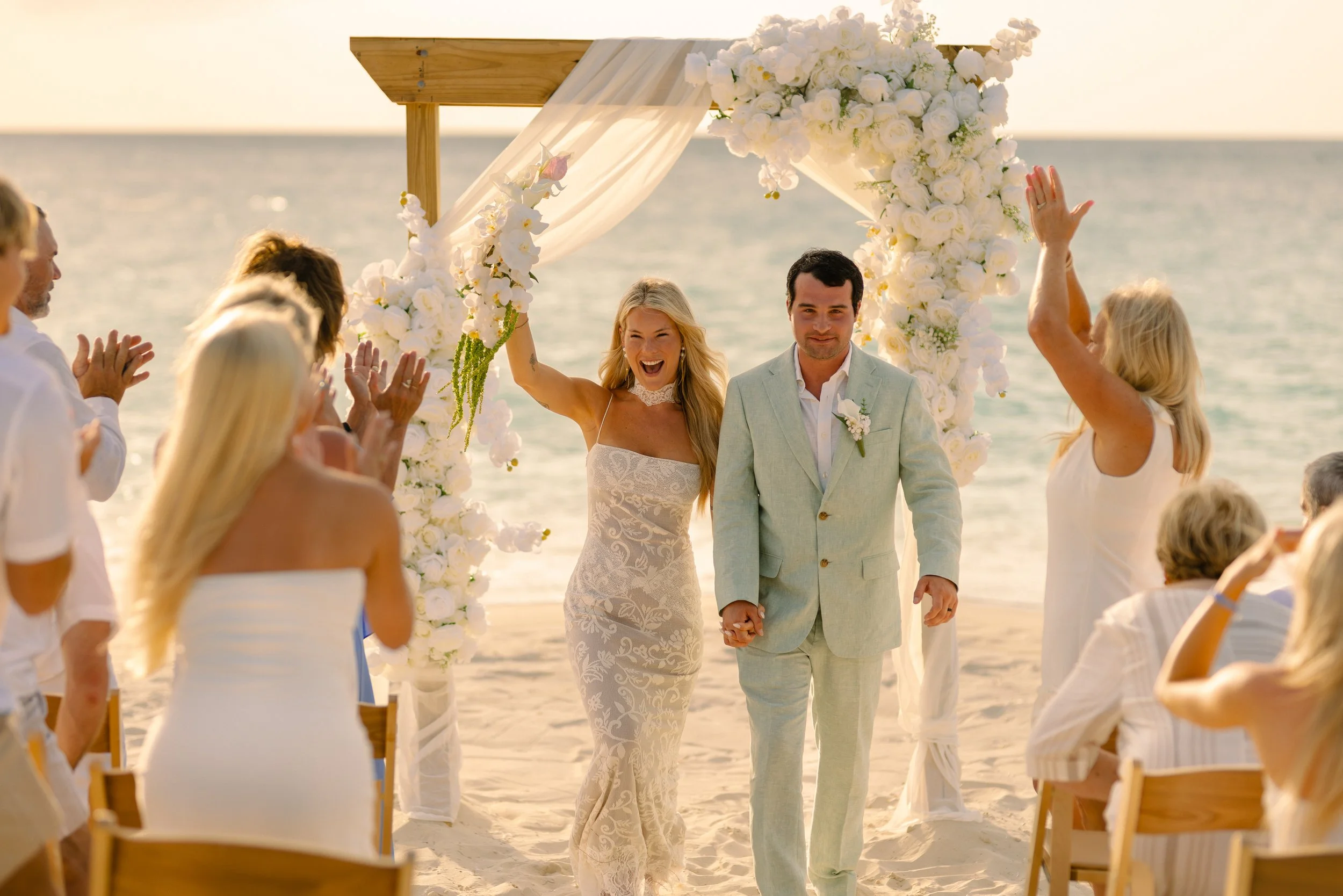 A bride and groom hold hands and walk through a beachside wedding ceremony arch decorated with white flowers, surrounded by seated guests clapping, with the ocean in the background during sunset.