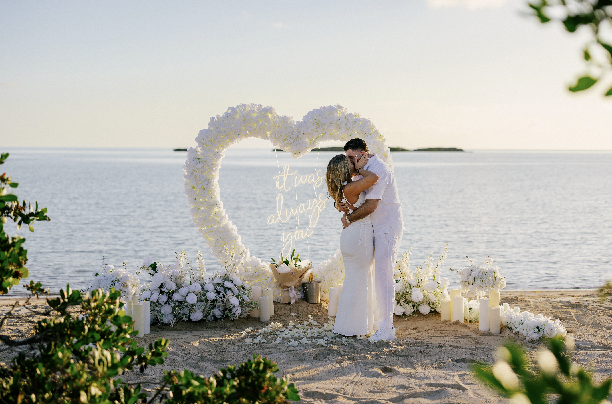 A couple kissing at a beach wedding ceremony with a heart-shaped floral arch, white flowers, candles, and a sign that reads 'it was always you' against the ocean backdrop.