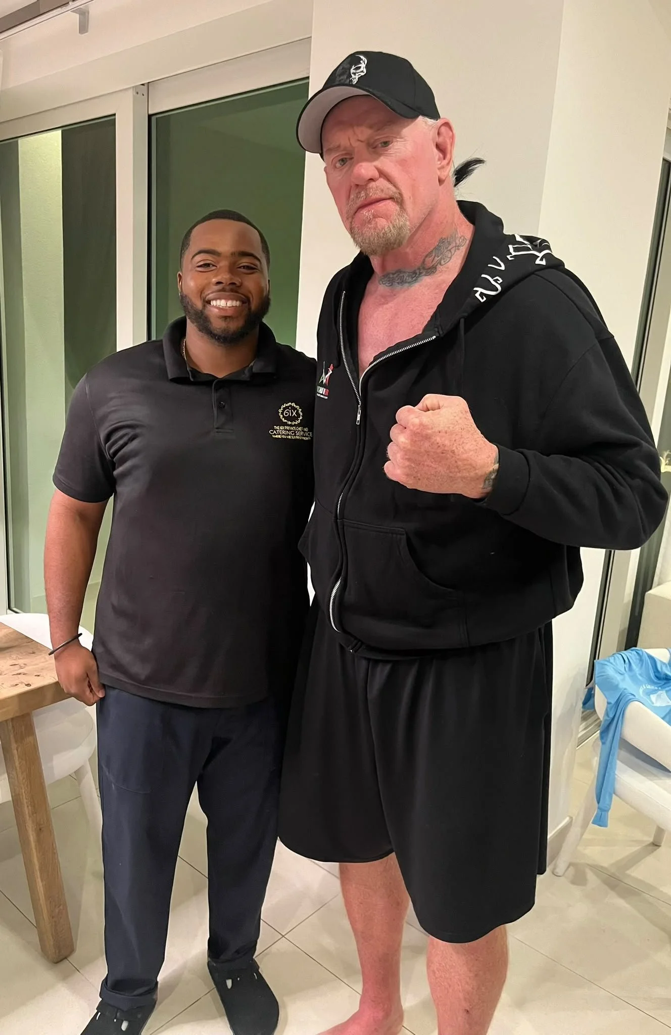 Two men standing together indoors, one in casual attire and the other shirtless in shorts and a hoodie, making a fist gesture, with guests in the background.