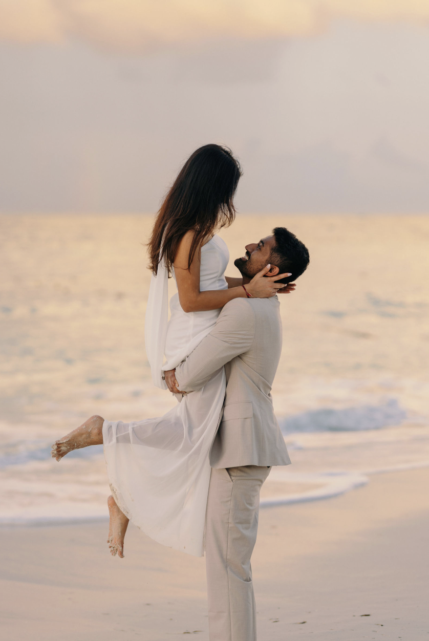A man in a beige suit lifting a woman in a white dress on a beach at sunset, with the ocean and sky in the background.