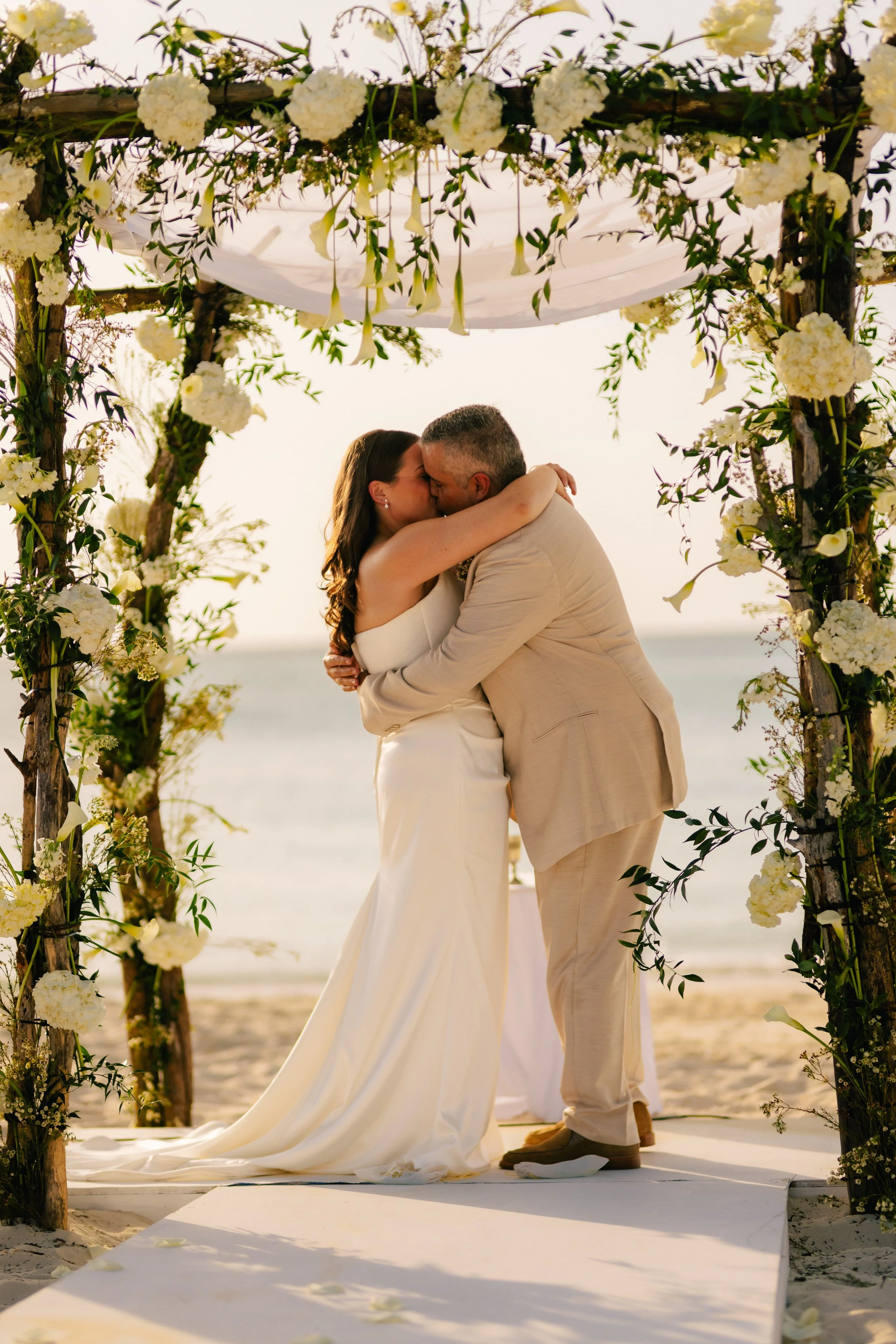 A couple sharing a kiss during their beach wedding ceremony, standing under a floral arch decorated with white flowers and greenery, with the ocean in the background at sunset.