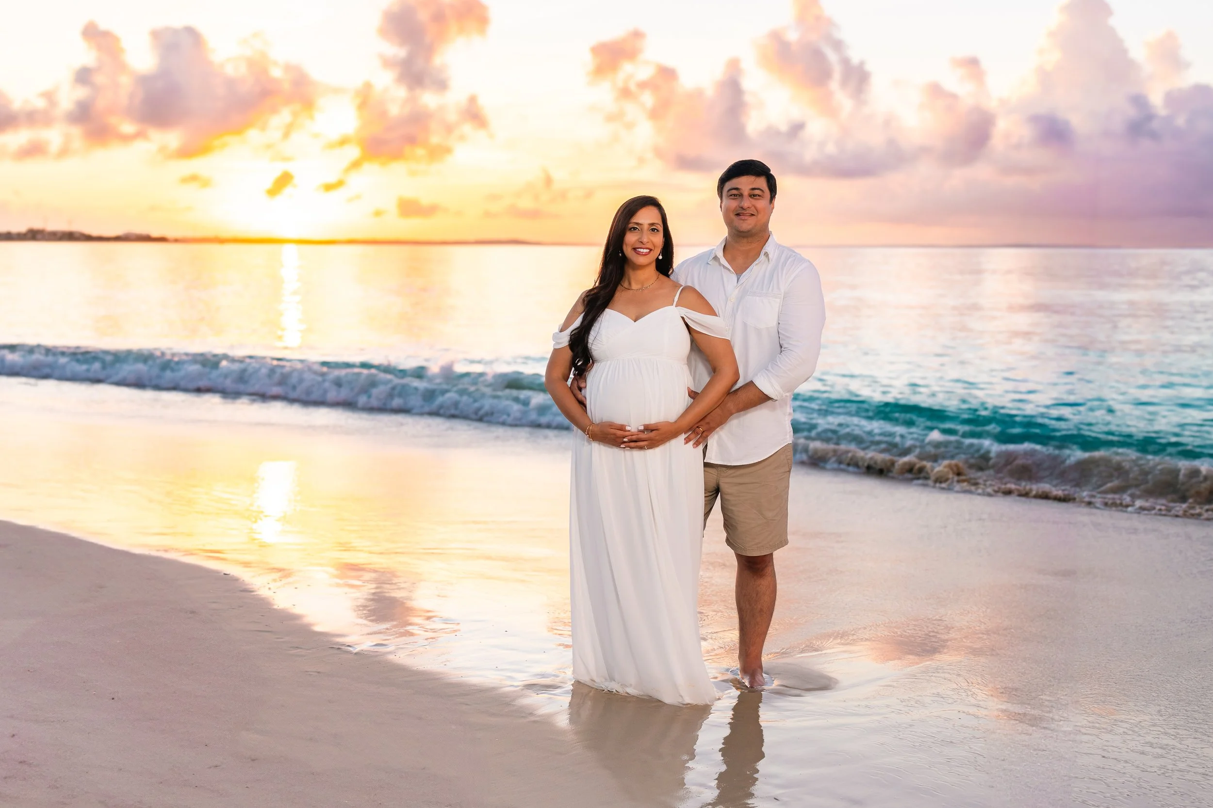 A couple standing on the beach during sunset, with the woman in a white dress and the man in a white shirt and khaki shorts, both smiling, with their hands on her pregnant belly, and the ocean and sky with clouds in the background.