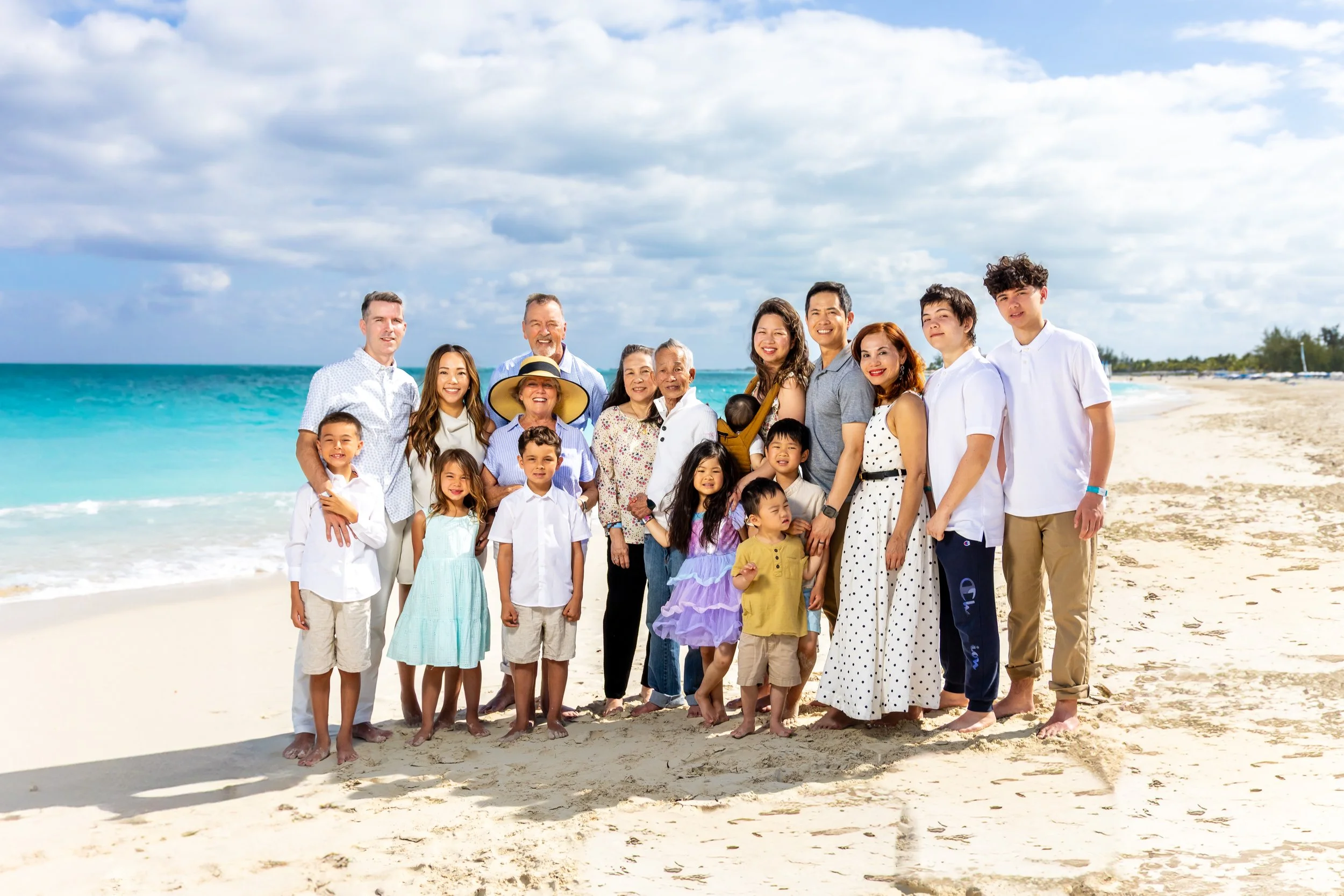 A large group of diverse people, including children and seniors, standing on the beach with the ocean and cloudy sky in the background.
