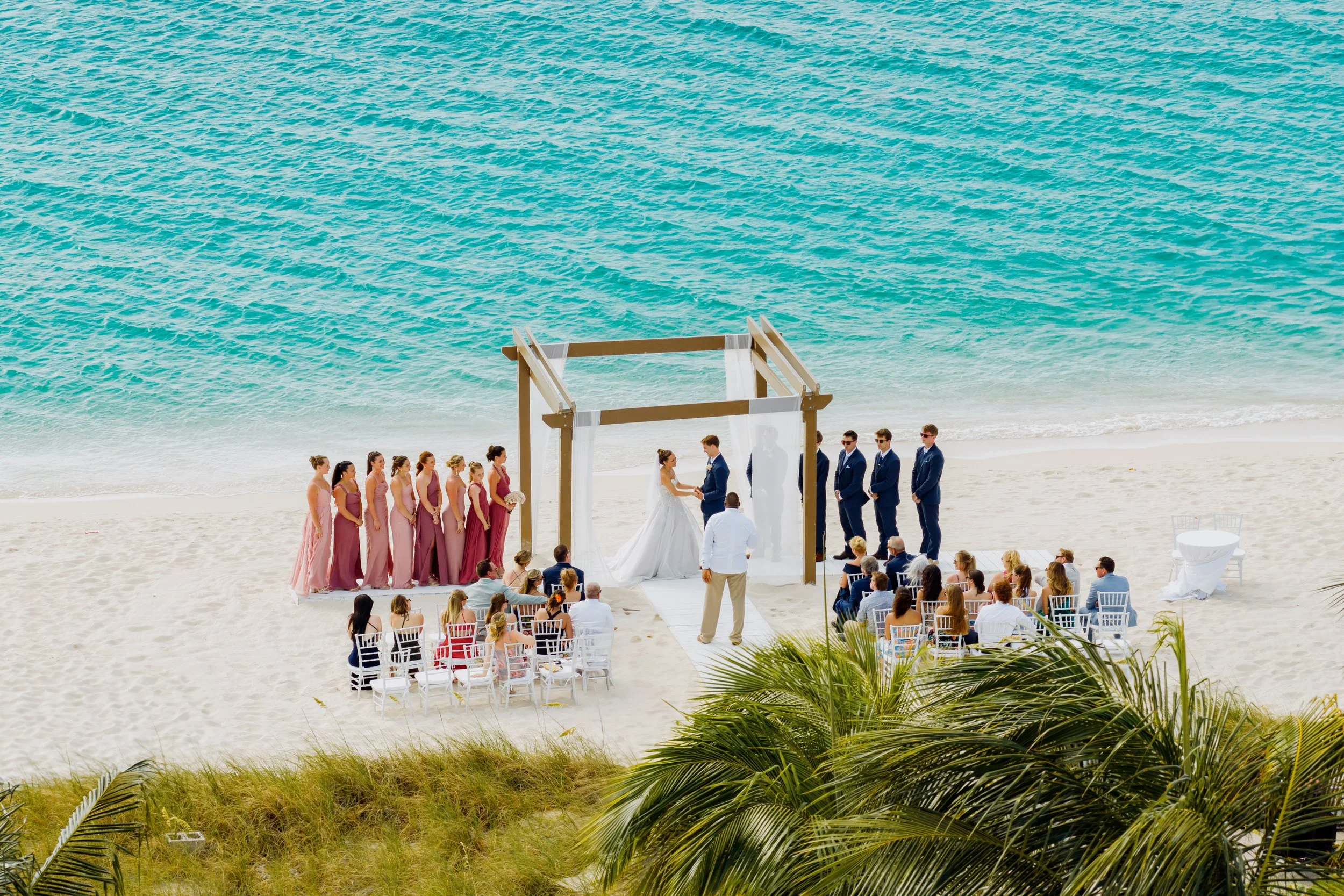 A beach wedding ceremony with a bride and groom exchanging vows under a wooden arch, surrounded by bridesmaids and groomsmen, audience seated on chairs facing the ocean, palm trees in the foreground, and turquoise water in the background.