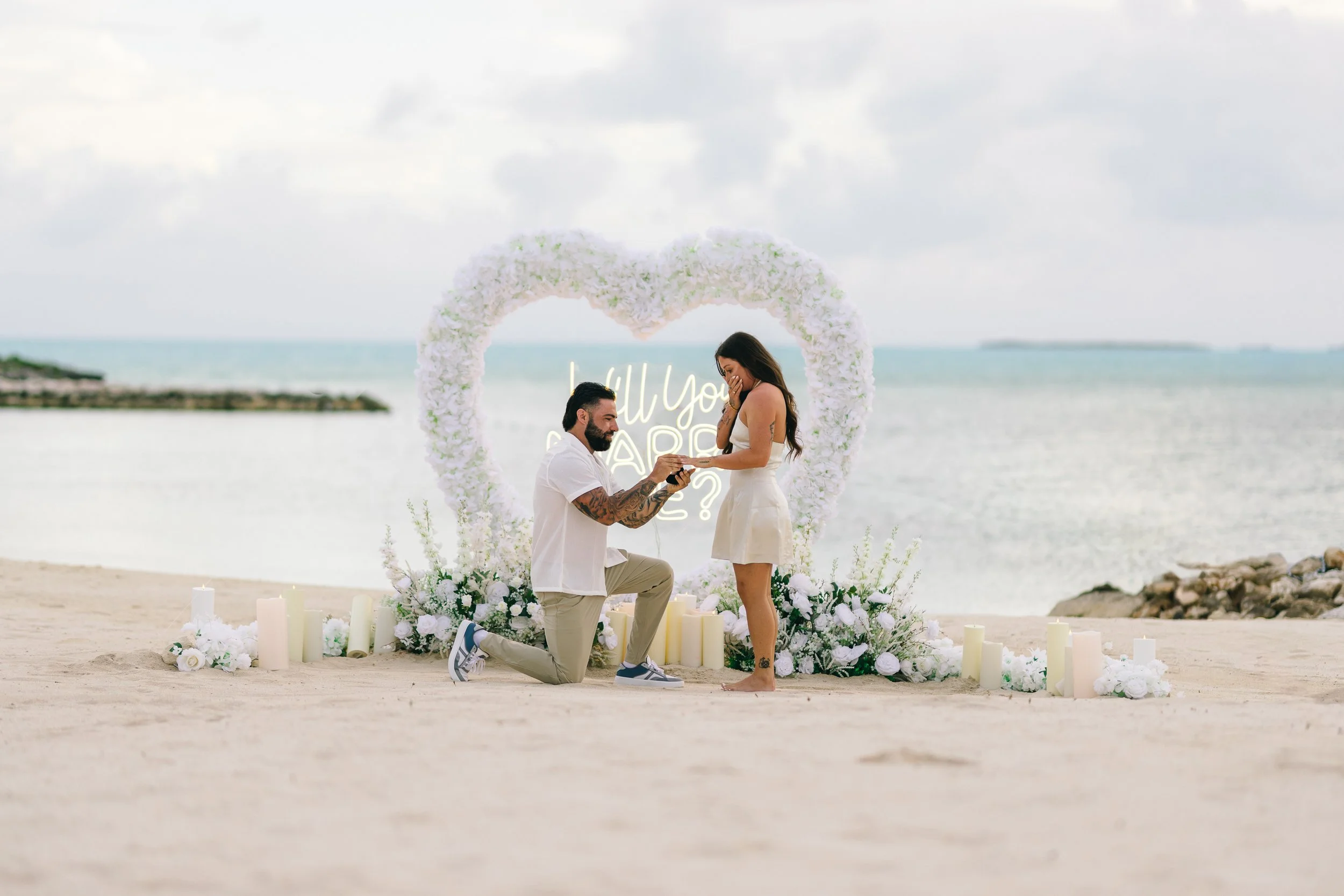 A man proposing marriage to a woman on a beach with a floral heart-shaped backdrop and candles, during daytime with cloudy sky.