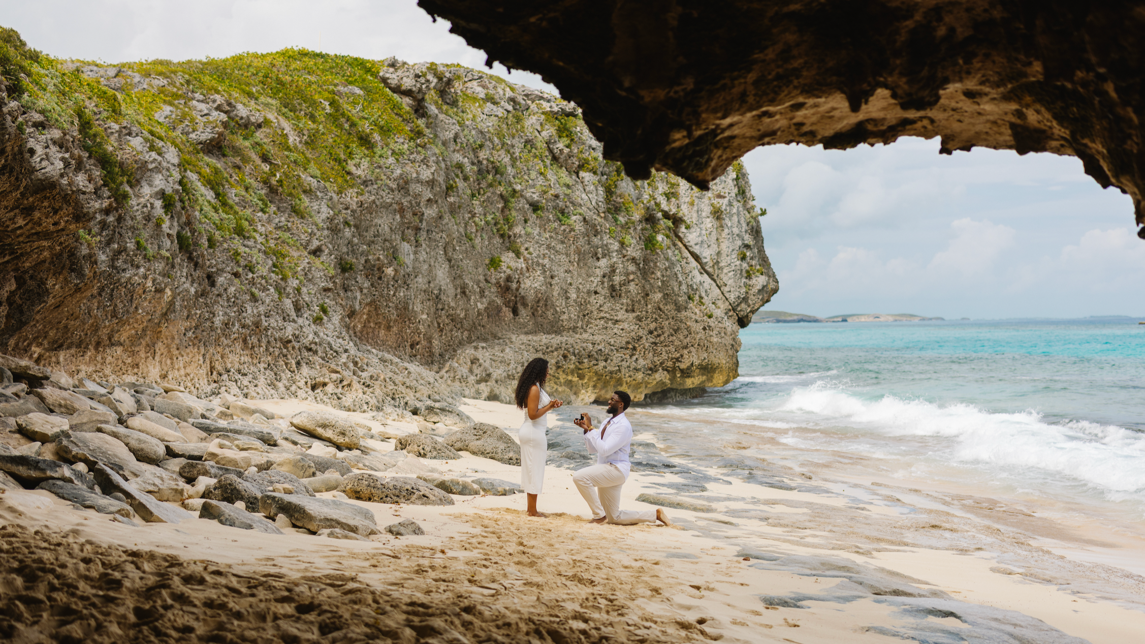 A couple gets engaged on a sandy beach with rocky shoreline, green cliffs, and ocean waves in the background.