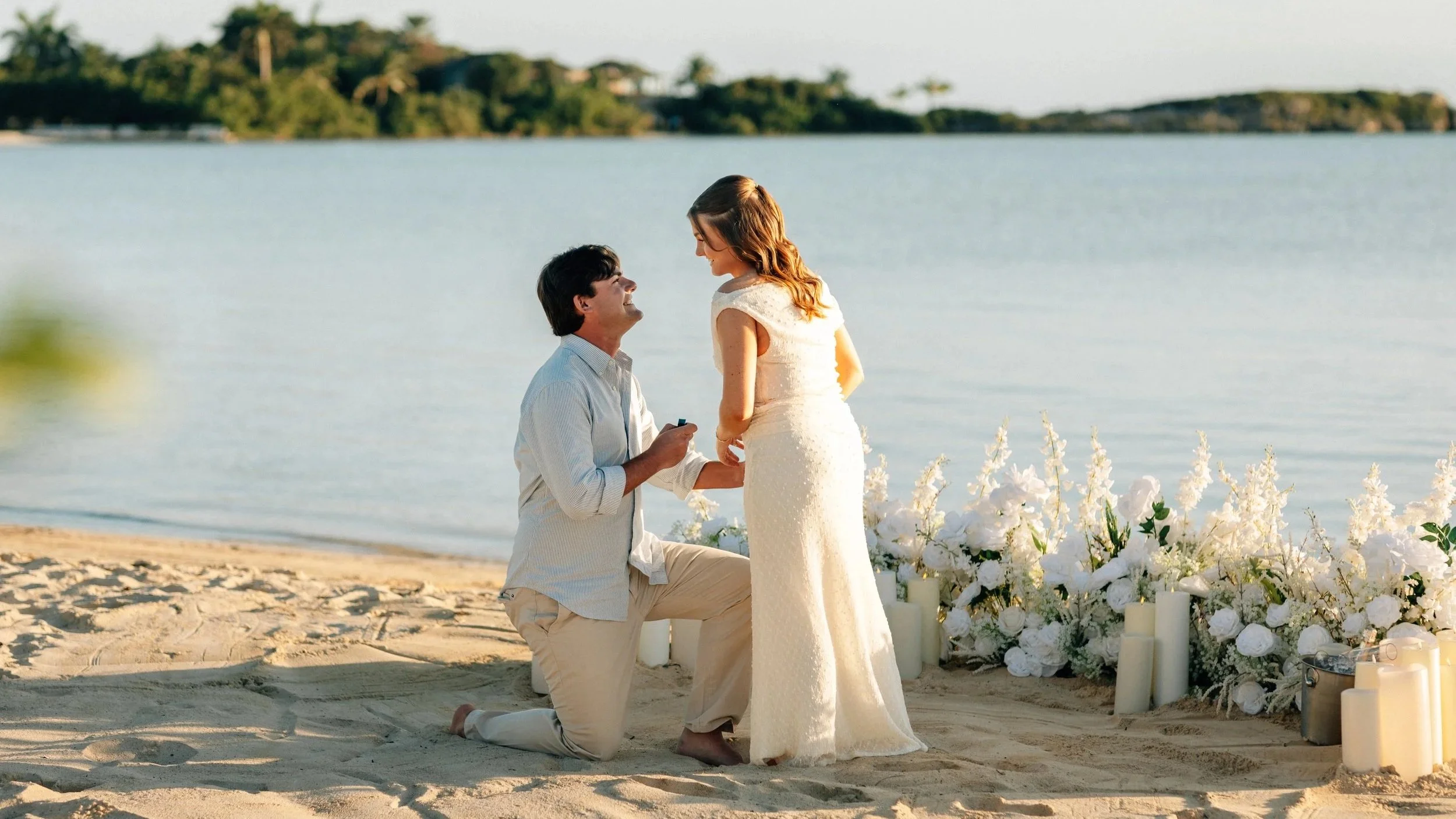 A couple participates in a wedding ceremony on a beach, with the man kneeling and proposing to the woman who is in a white wedding dress, surrounded by white flowers and candles by the water.