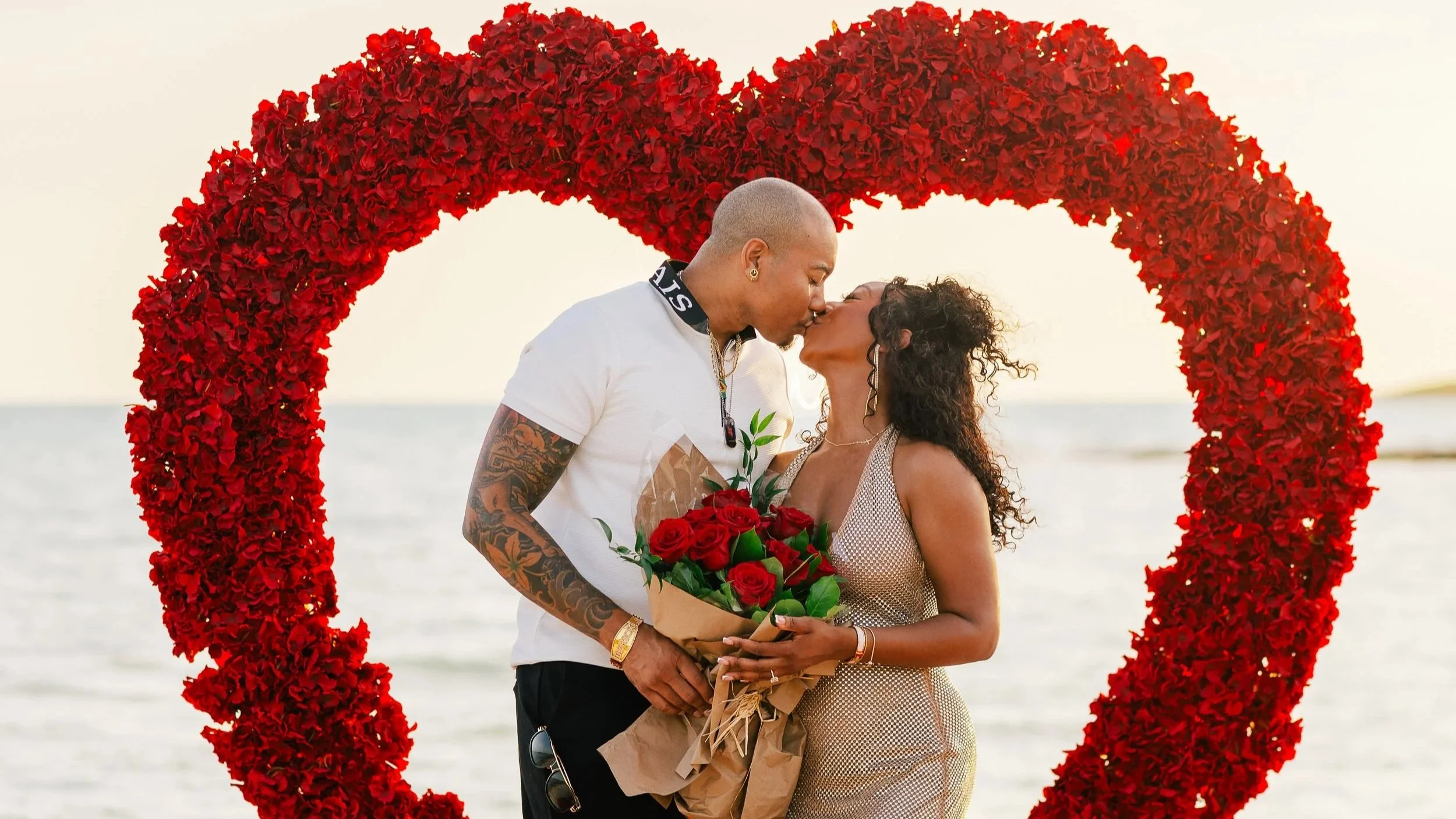 A couple sharing a kiss in front of a heart-shaped flower arch with the ocean in the background.