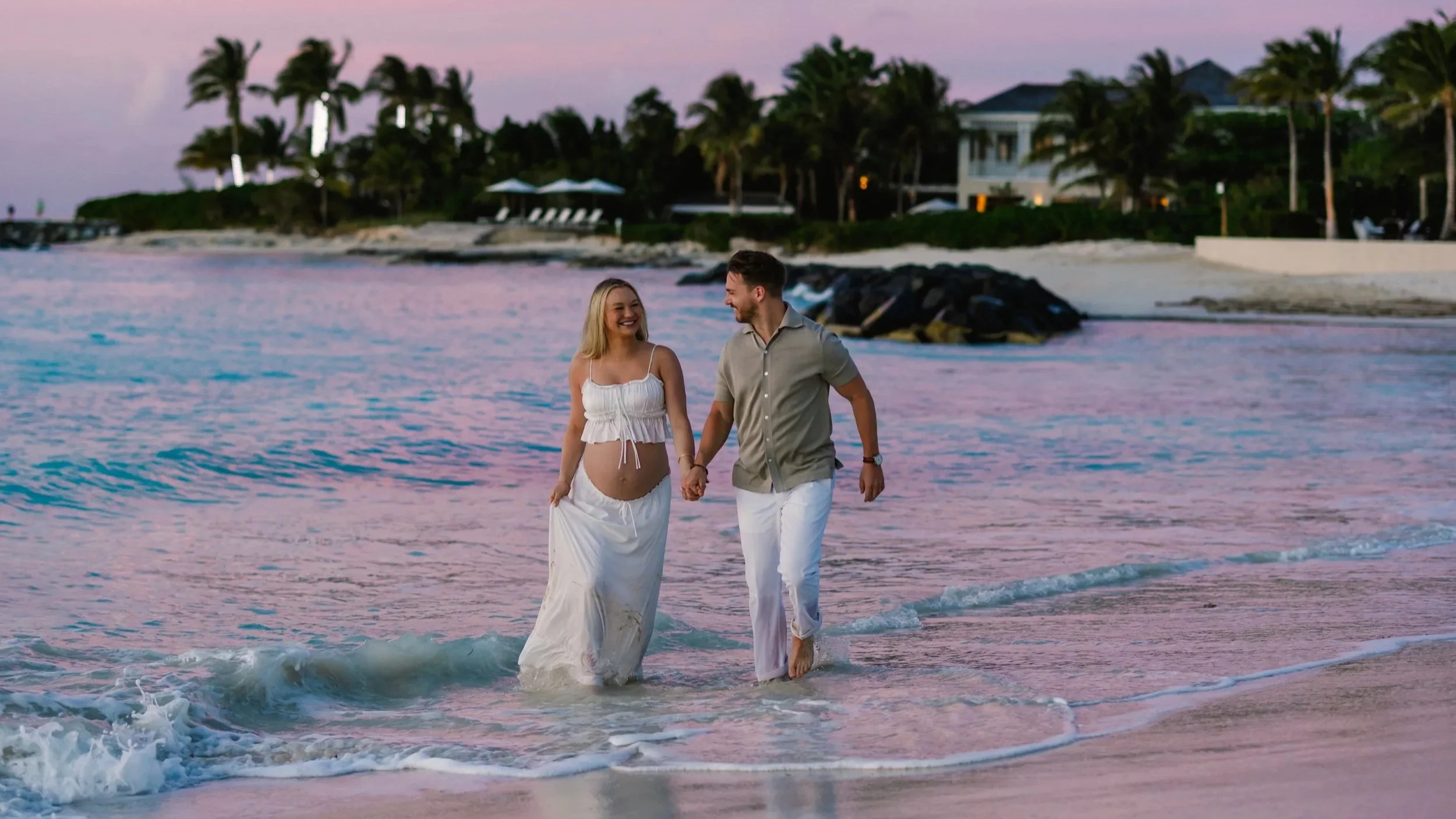 A smiling woman and man walking hand-in-hand along the shoreline at sunset, with the woman wearing a white dress and the man in light-colored clothes, near a beach with palm trees and beachfront houses in the background.