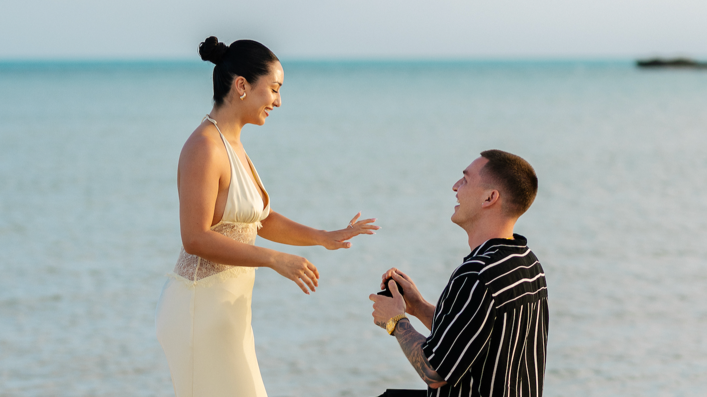 A woman in a yellow dress is proposing to a man on the beach, with the ocean in the background. The man is kneeling and holding a ring, smiling up at her.