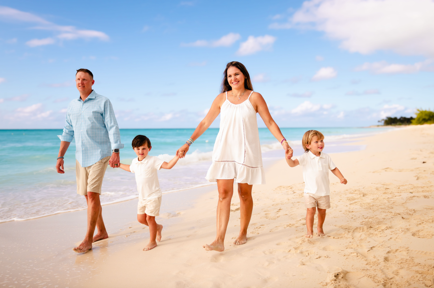 A family of four walking on a sandy beach near the ocean, holding hands and smiling. The adult woman is in the center, wearing a white summer dress and the adult man is on the left, wearing a light blue shirt and beige shorts. The two children, boys,