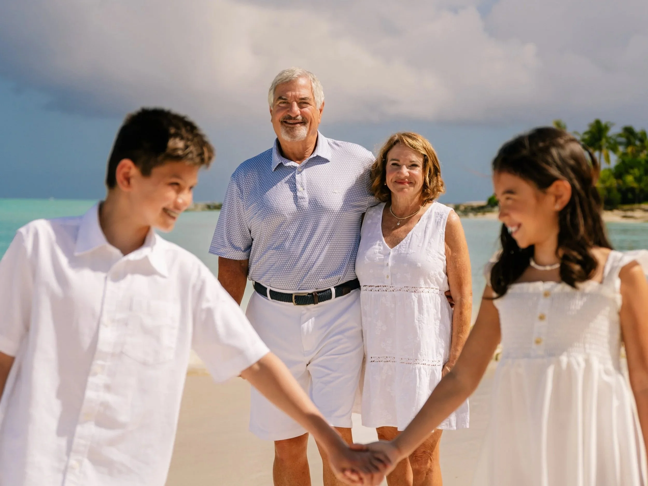 Family of four holding hands on the beach, with cloudy sky and palm trees in the background.