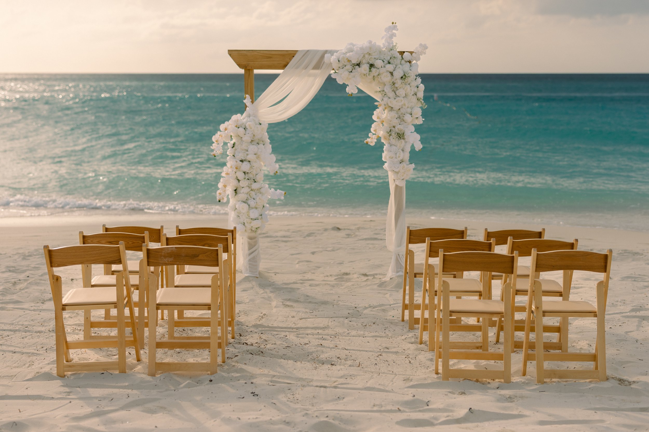 A wedding ceremony setup on a beach with wooden chairs arranged facing a floral arch draped with white fabric, overlooking the ocean with a cloudy sky.