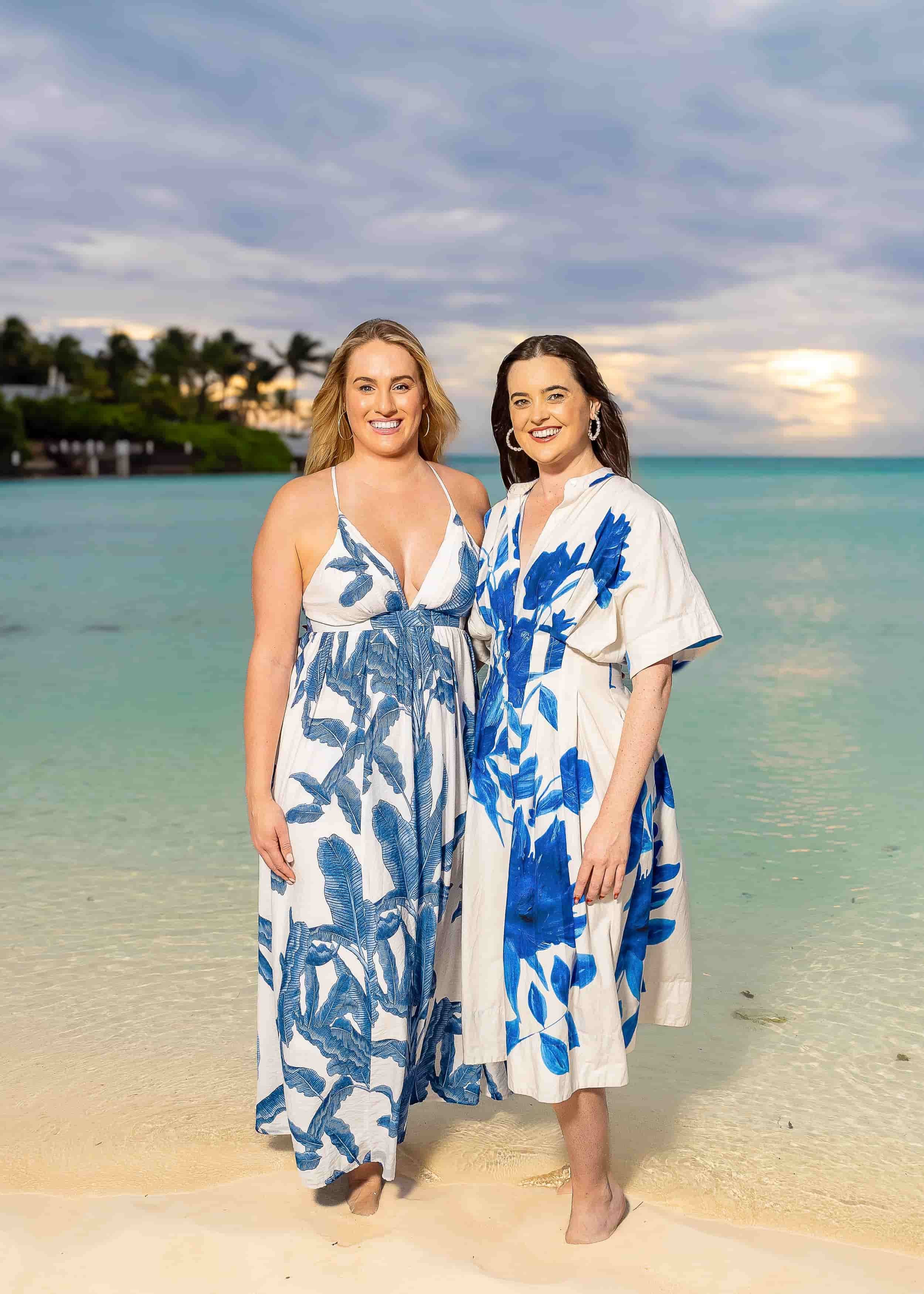 Two women standing on a beach with ocean and sky in the background, both wearing white dresses with blue floral patterns, smiling at the camera.