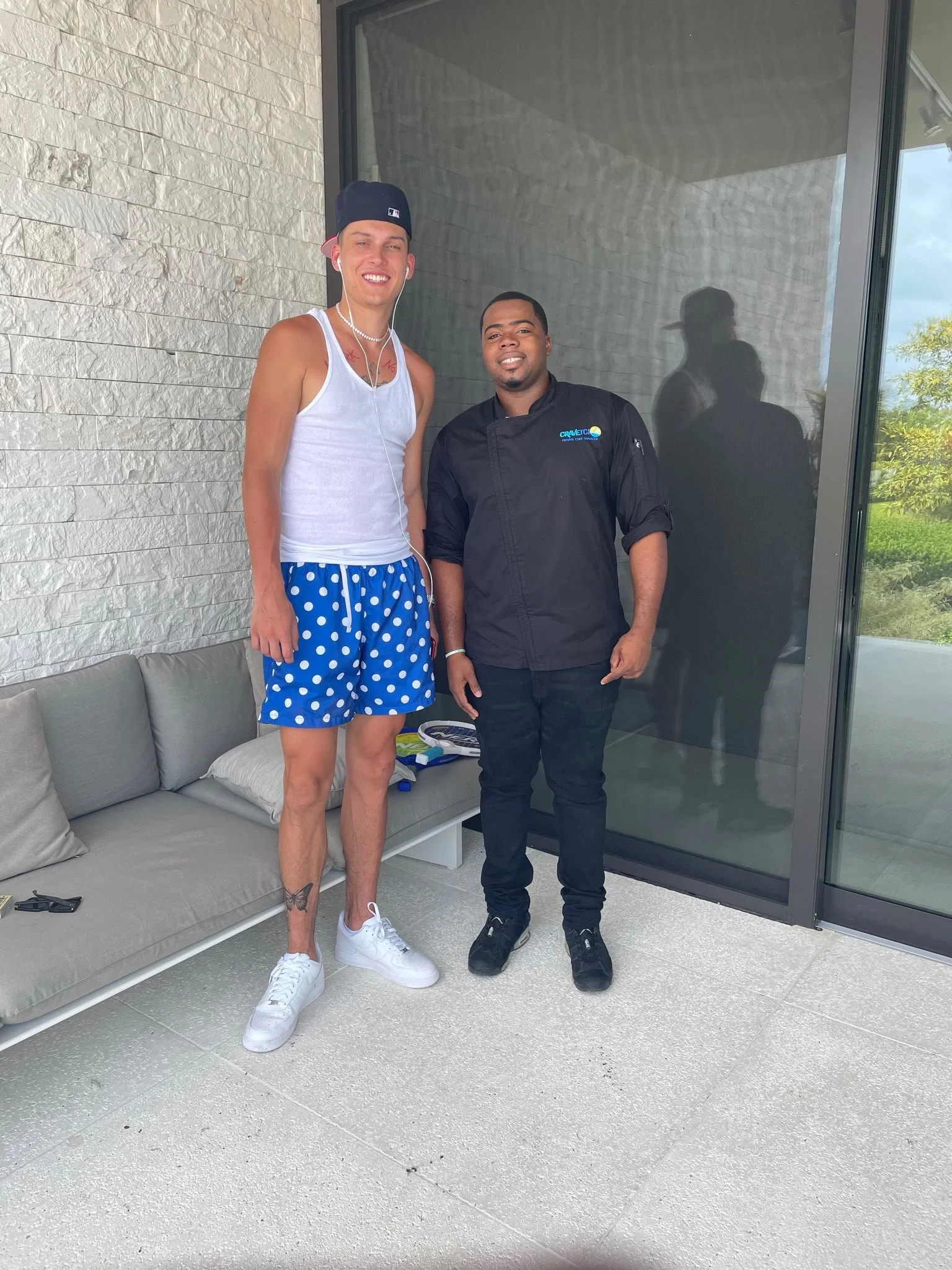 Two men standing outside near a glass door and sitting area. One is wearing a white tank top, blue polka dot shorts, white sneakers, a cap worn backward, and earbuds. The other is wearing a black chef's coat and black shoes. The background has a white textured wall and green trees.