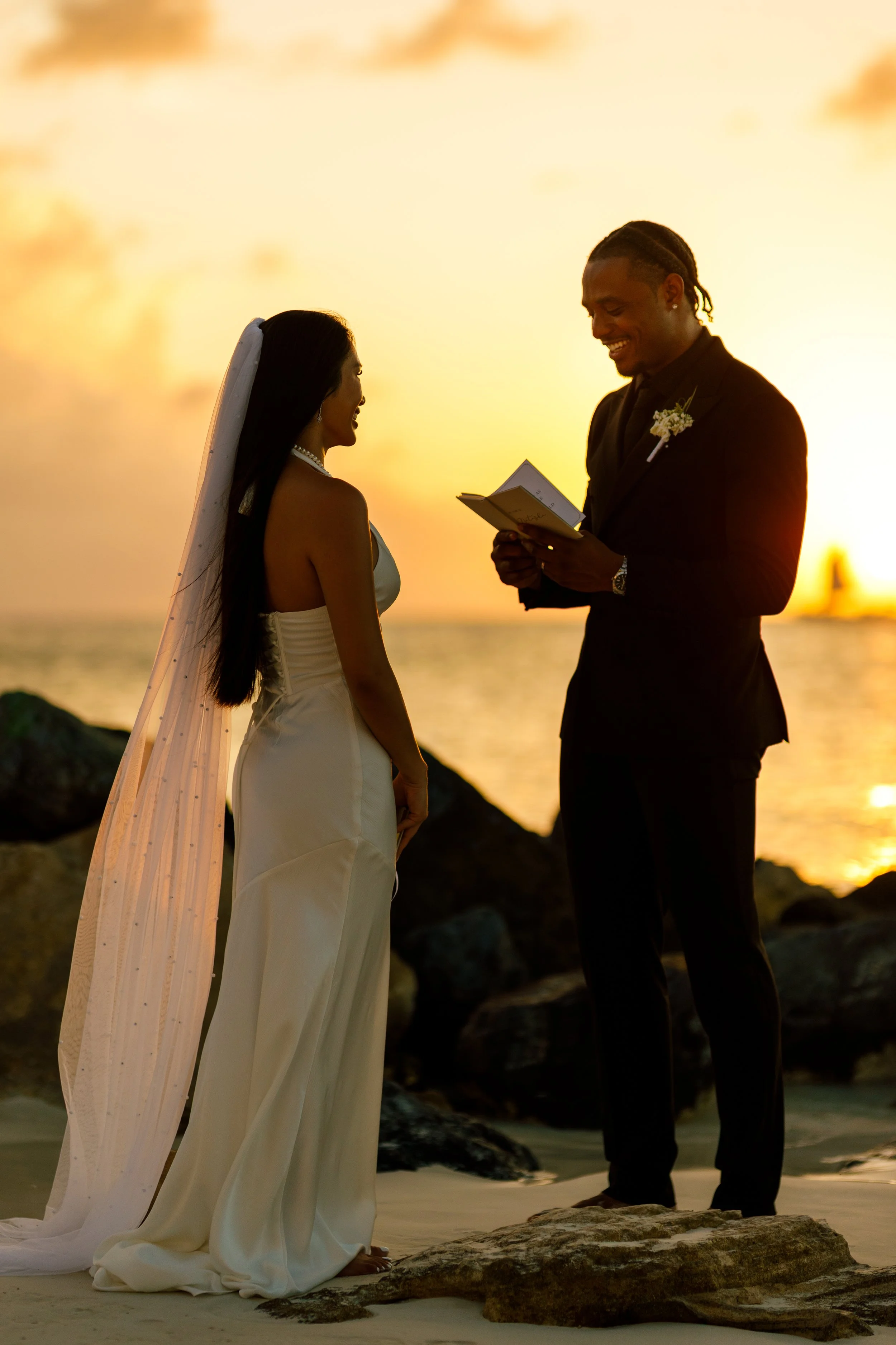 A bride and groom exchanging vows on the beach at sunset with rocks and water in the background.