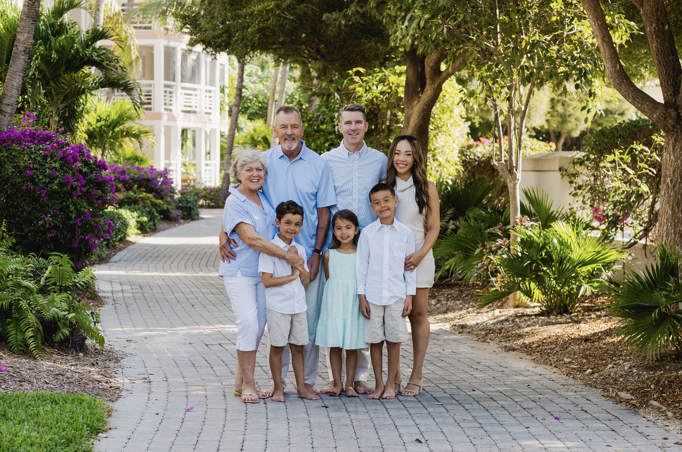 A multigenerational family of nine people, including children and older adults, posing together outdoors on a stone pathway surrounded by lush green tropical plants and trees on a sunny day.