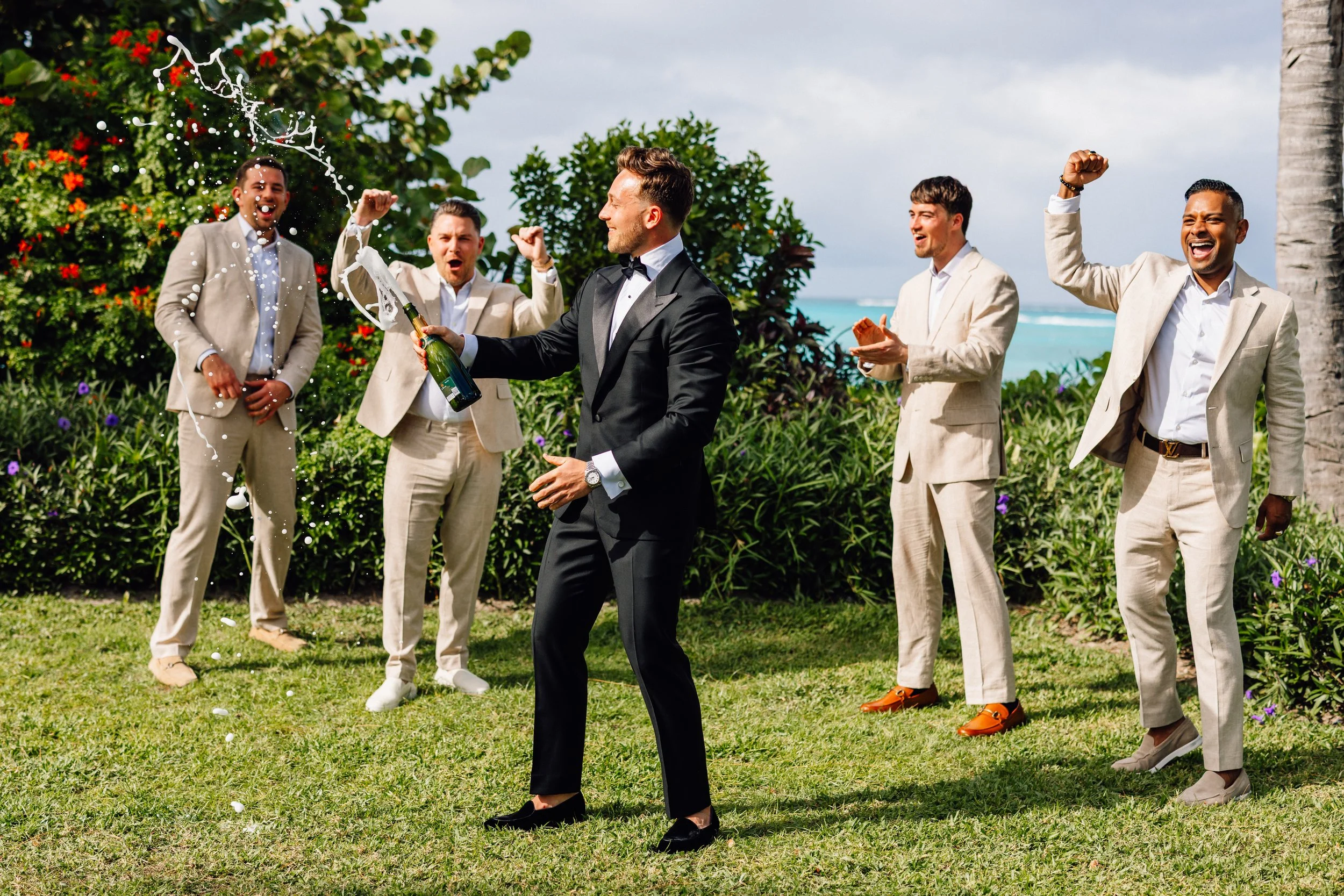 A group of five men in suits celebrating outdoors on a grassy area next to the beach, with one man in a black tuxedo popping a champagne bottle, creating a splash, while the others cheer and smile. The background features tropical plants, trees, and 