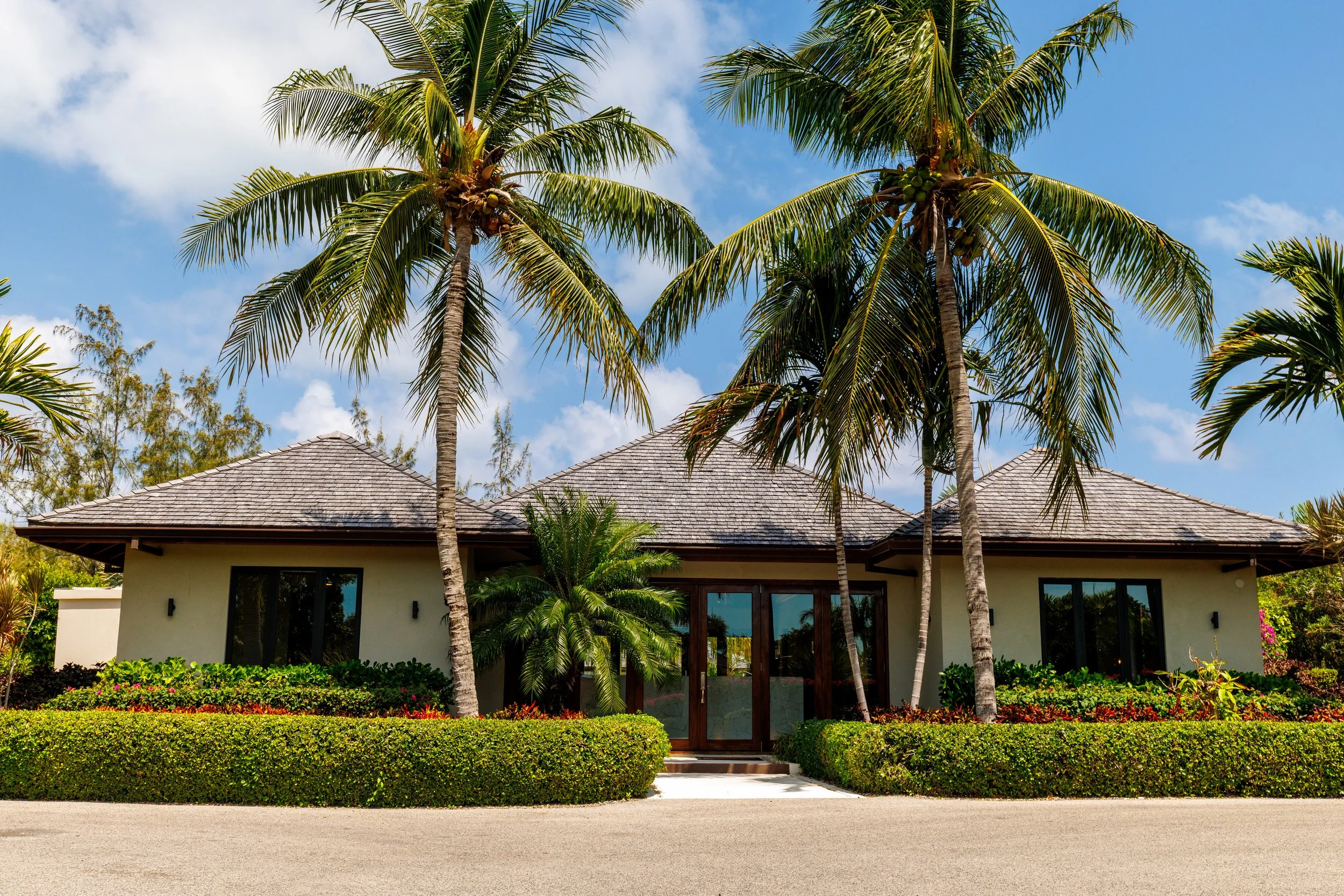 Modern house with tropical landscaping featuring tall palm trees, a well-kept lawn, and a driveway in the foreground under a partly cloudy sky.
