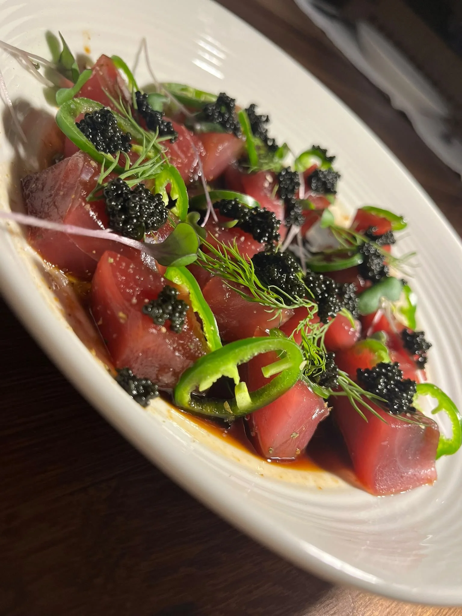 Crispy tuna poke with slices of raw tuna, topped with black caviar, green onion, herbs, and microgreens, served in a white dish.