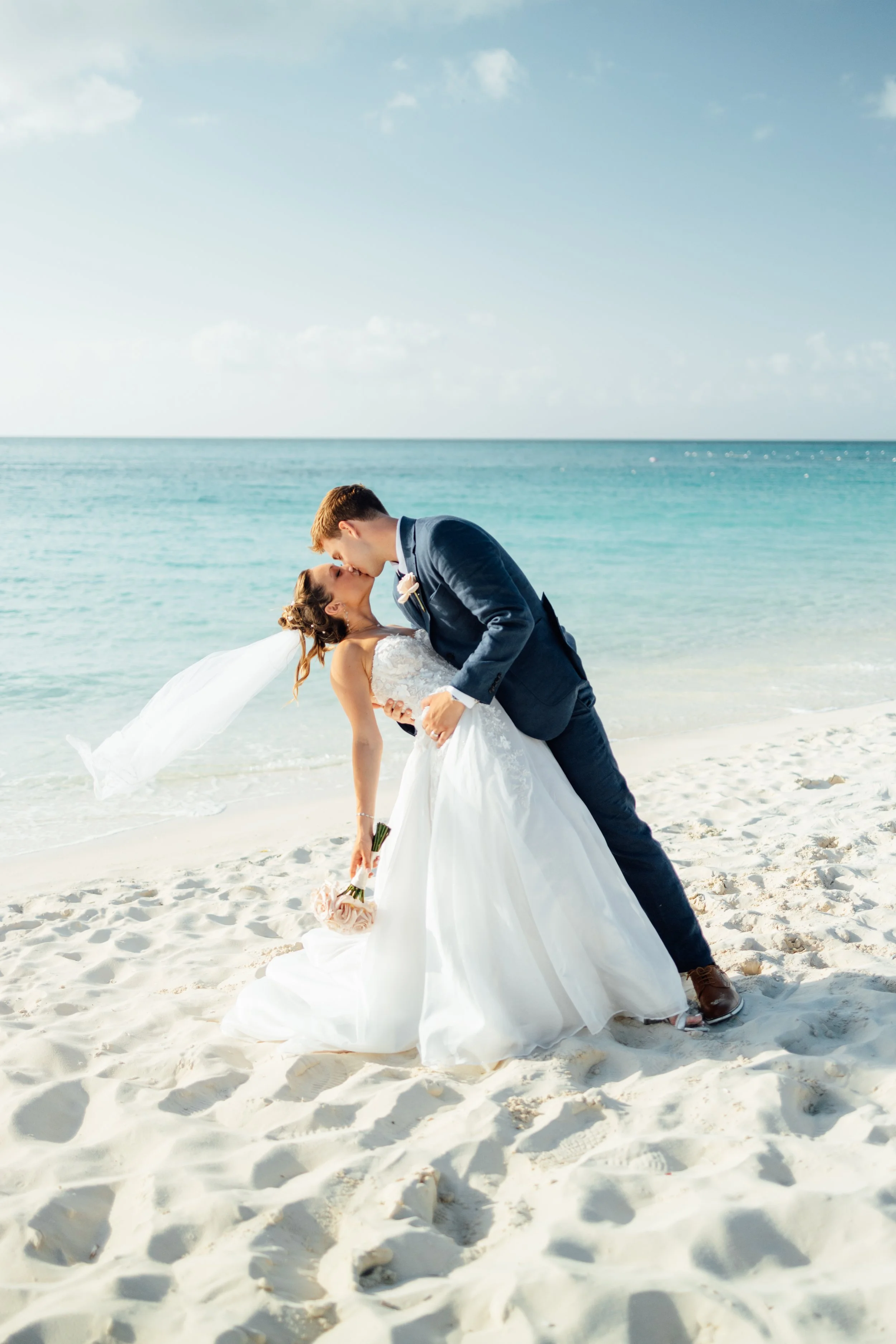 A newlywed couple is dancing on the beach. The groom, wearing a dark suit, is dipping the bride, who is holding a bouquet of flowers. The bride is wearing a white wedding dress and veil. The ocean and sky are in the background.