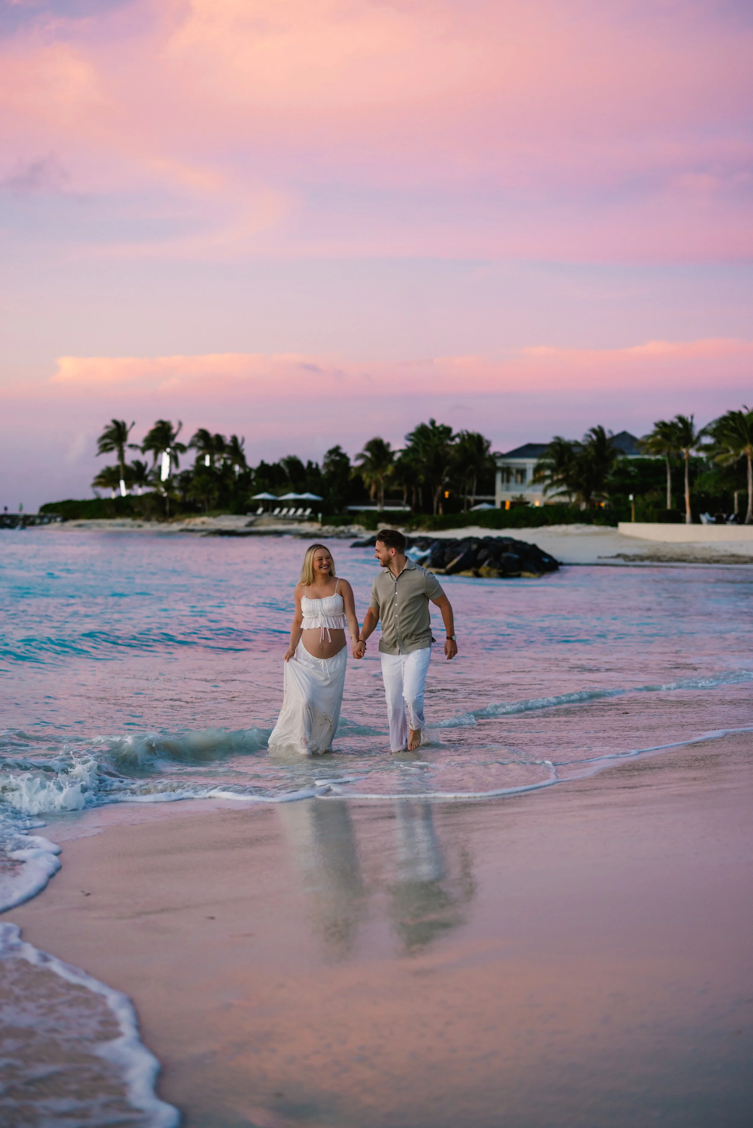 A couple walking hand in hand on the beach during sunset, with pink and purple skies and houses surrounded by palm trees in the background.