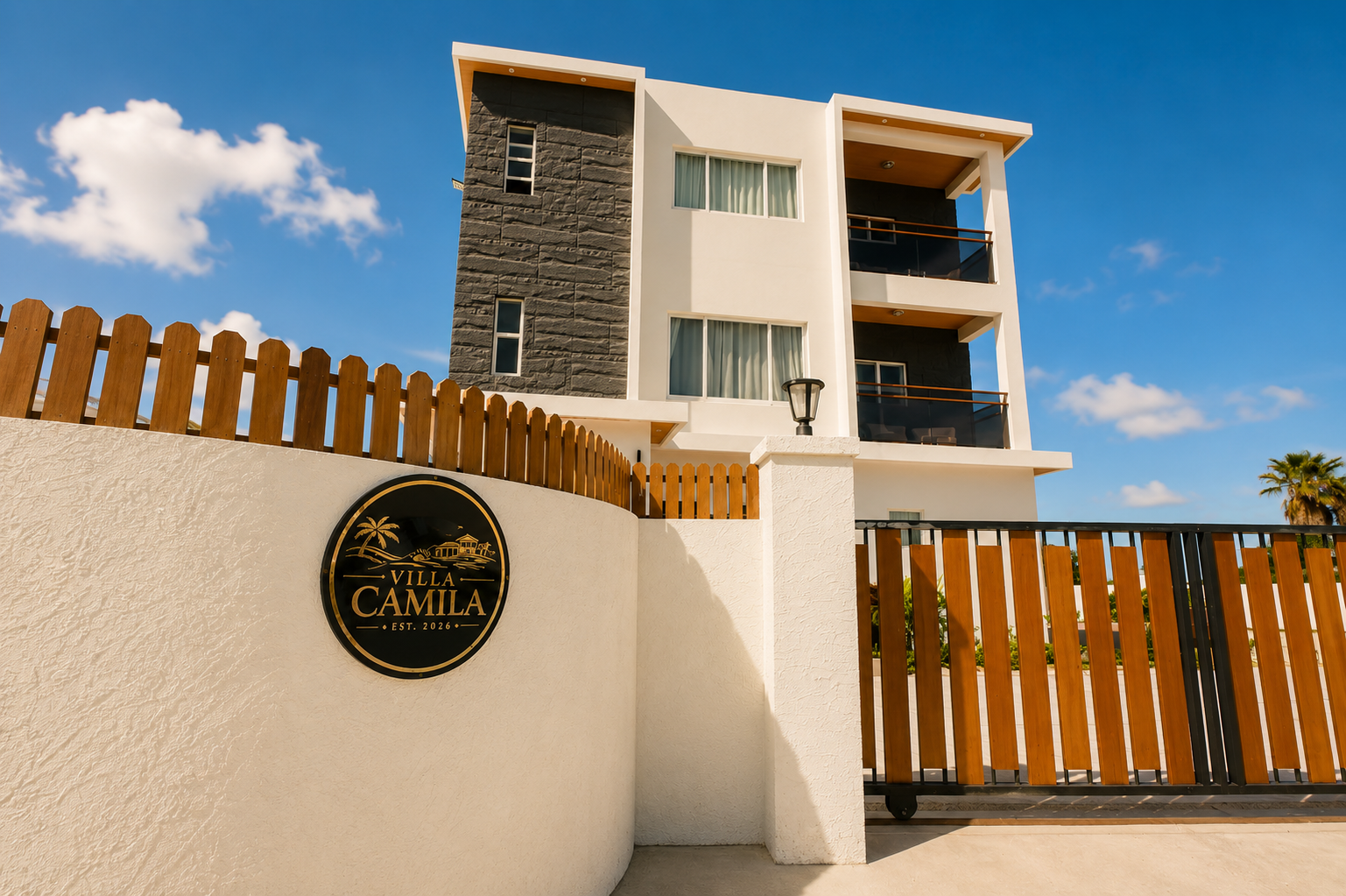 A modern multi-story villa with white and dark gray exterior walls, large windows with curtains, and balconies, surrounded by a white wall and wooden fencing, under a blue sky with clouds. A sign on the wall reads "Villa Camila, Est. 2026."